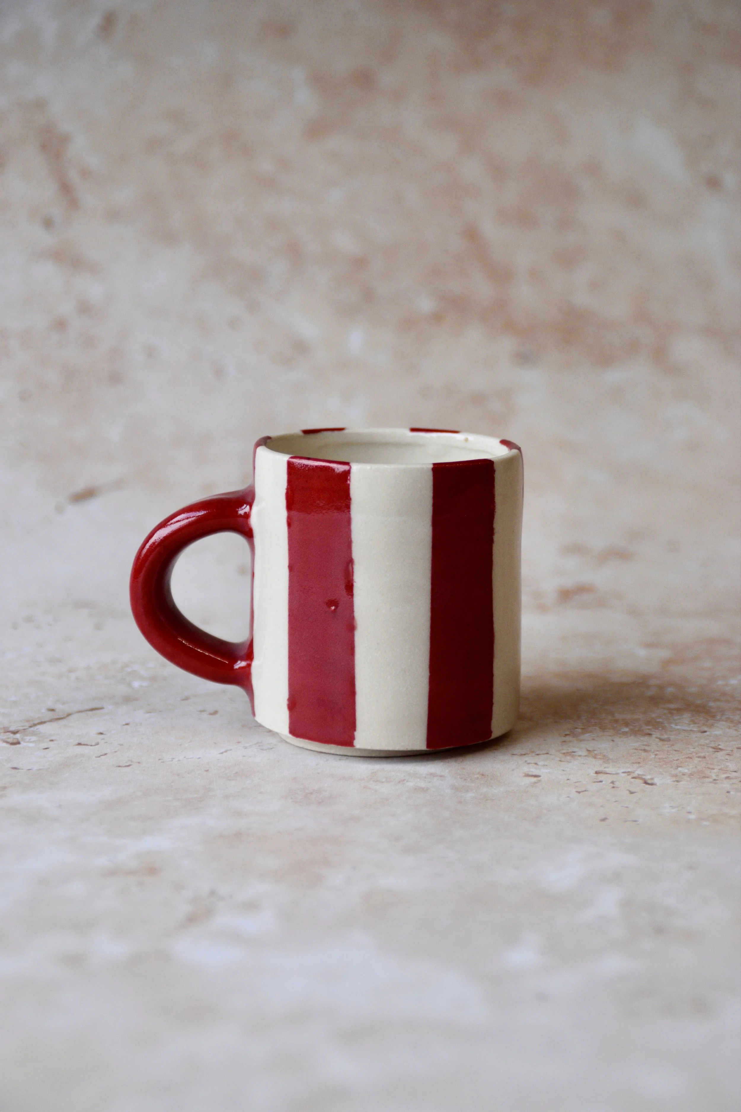 Handmade ceramic espresso cup with bold vertical red and cream stripes and glossy red handle, photographed on neutral background.