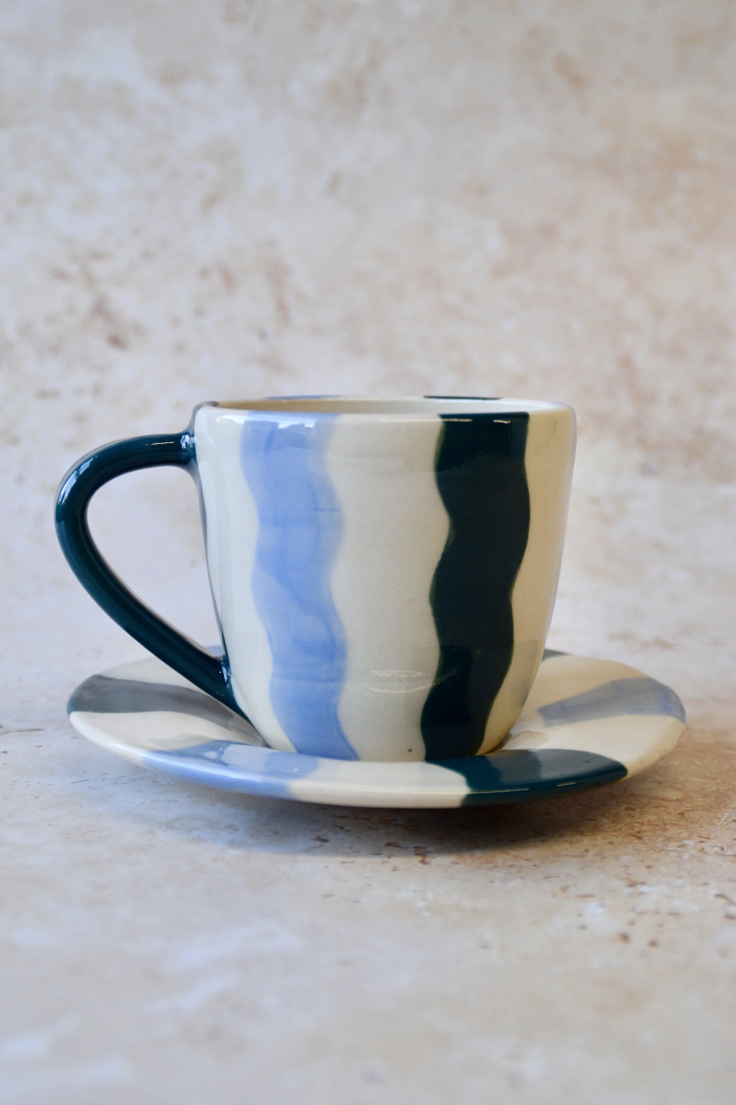 Handmade wavy striped ceramic cup and saucer in blue tones on a neutral background.