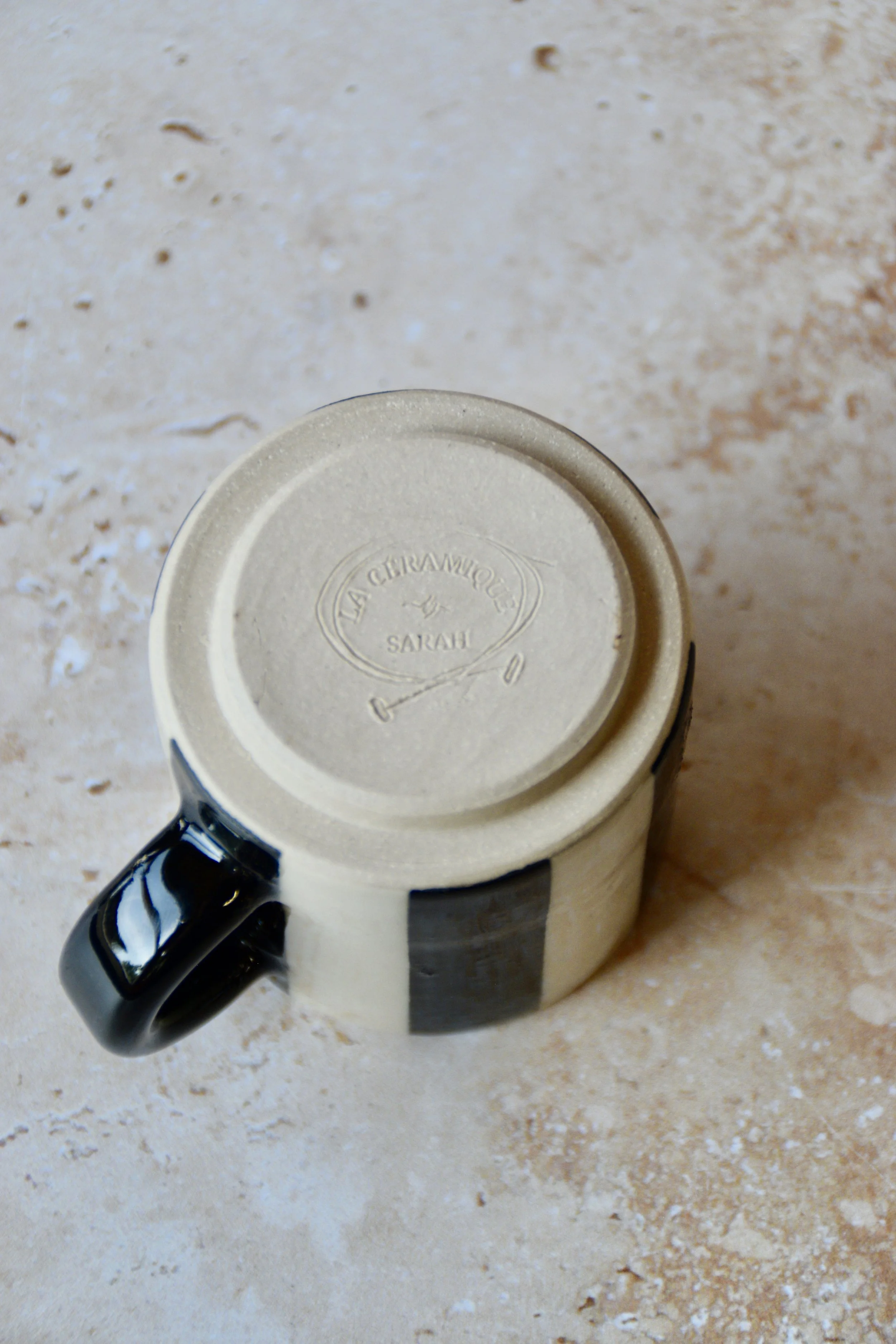 A ceramic mug with a black handle and a cream-coloured body. The mug features vertical black stripes and the text "LA CERAMIQUE by SARAH" stamped on the bottom. The mug is shown upside down.