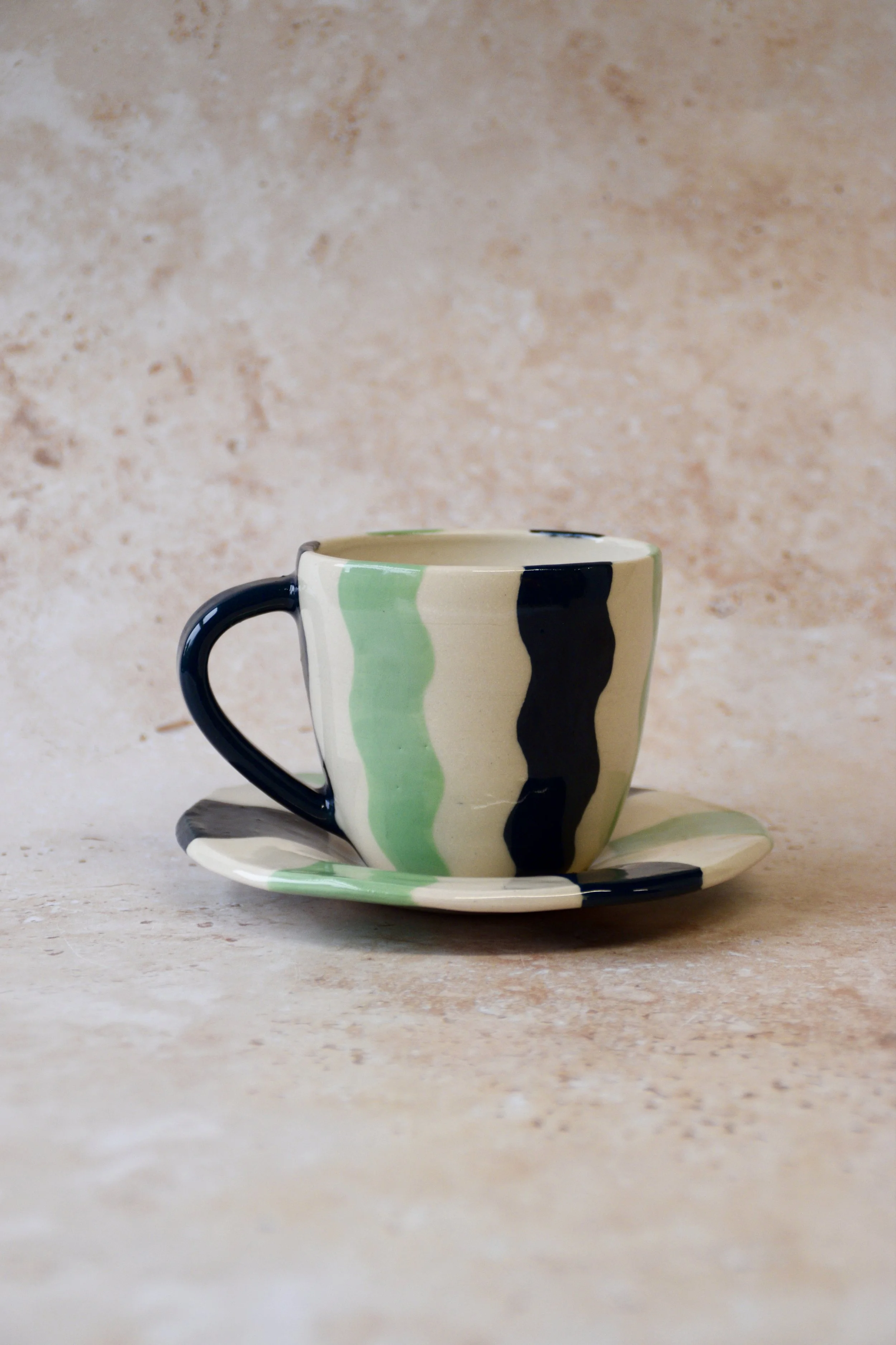 Handmade ceramic espresso cup and saucer with wavy stripe pattern in deep green and soft matcha tones on a cream base, photographed against a neutral textured background.