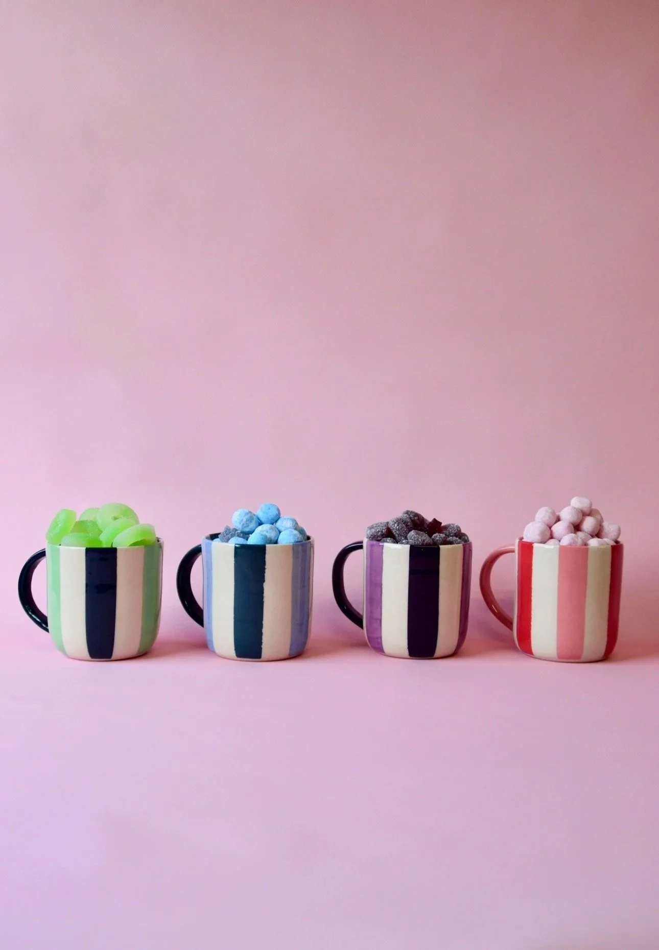Handmade striped ceramic mugs in four colourways – green, blue, purple and pink – filled with sweets and styled against a soft pink background.