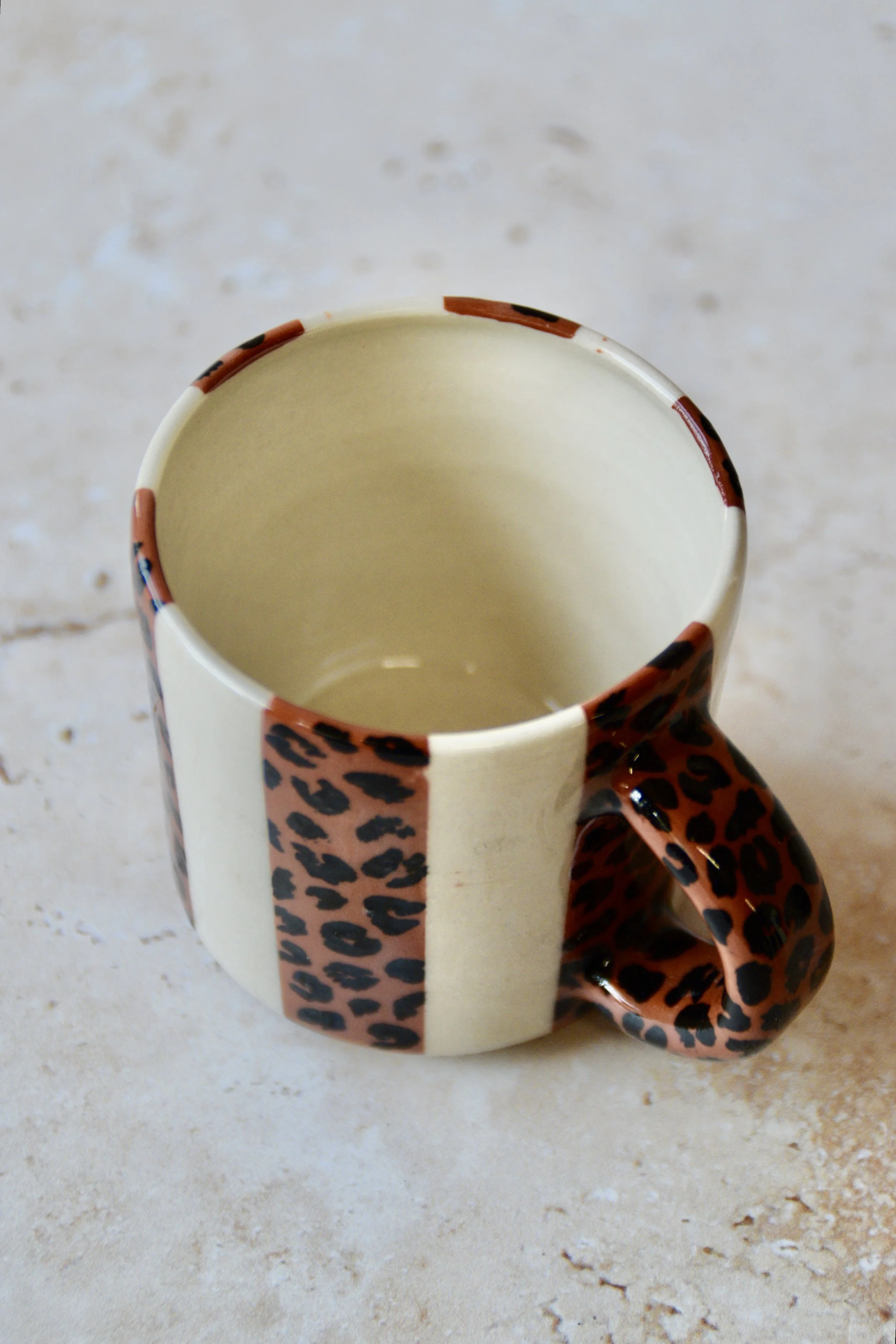 A ceramic mug with a cream interior and exterior. The exterior has alternating leopard and cream vertical stripes. The handle is leopard print. The mug is designed for hot drinks.
