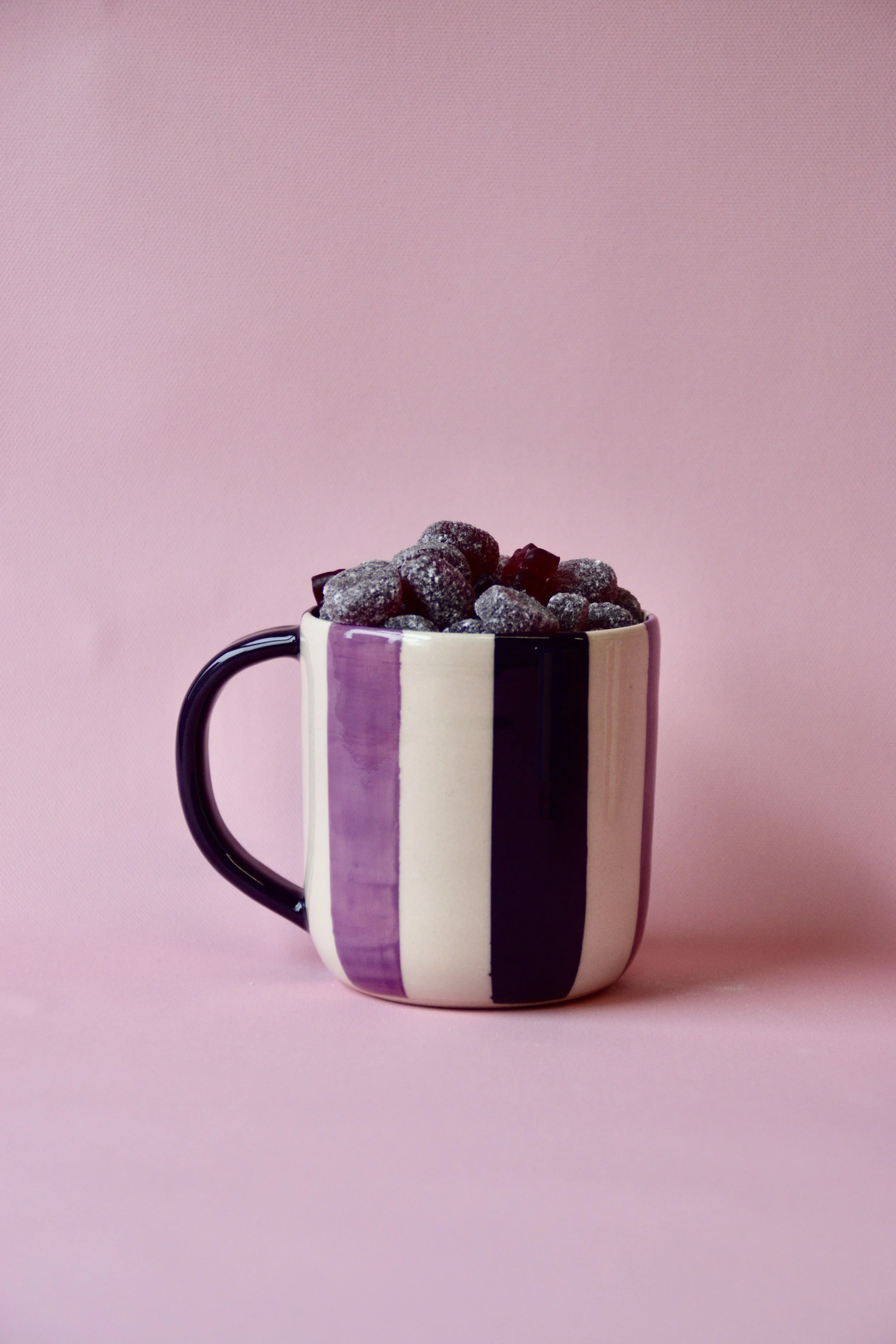 Handmade purple and cream striped ceramic mug with dark plum handle, filled with dark sugared sweets, styled on a pink background.