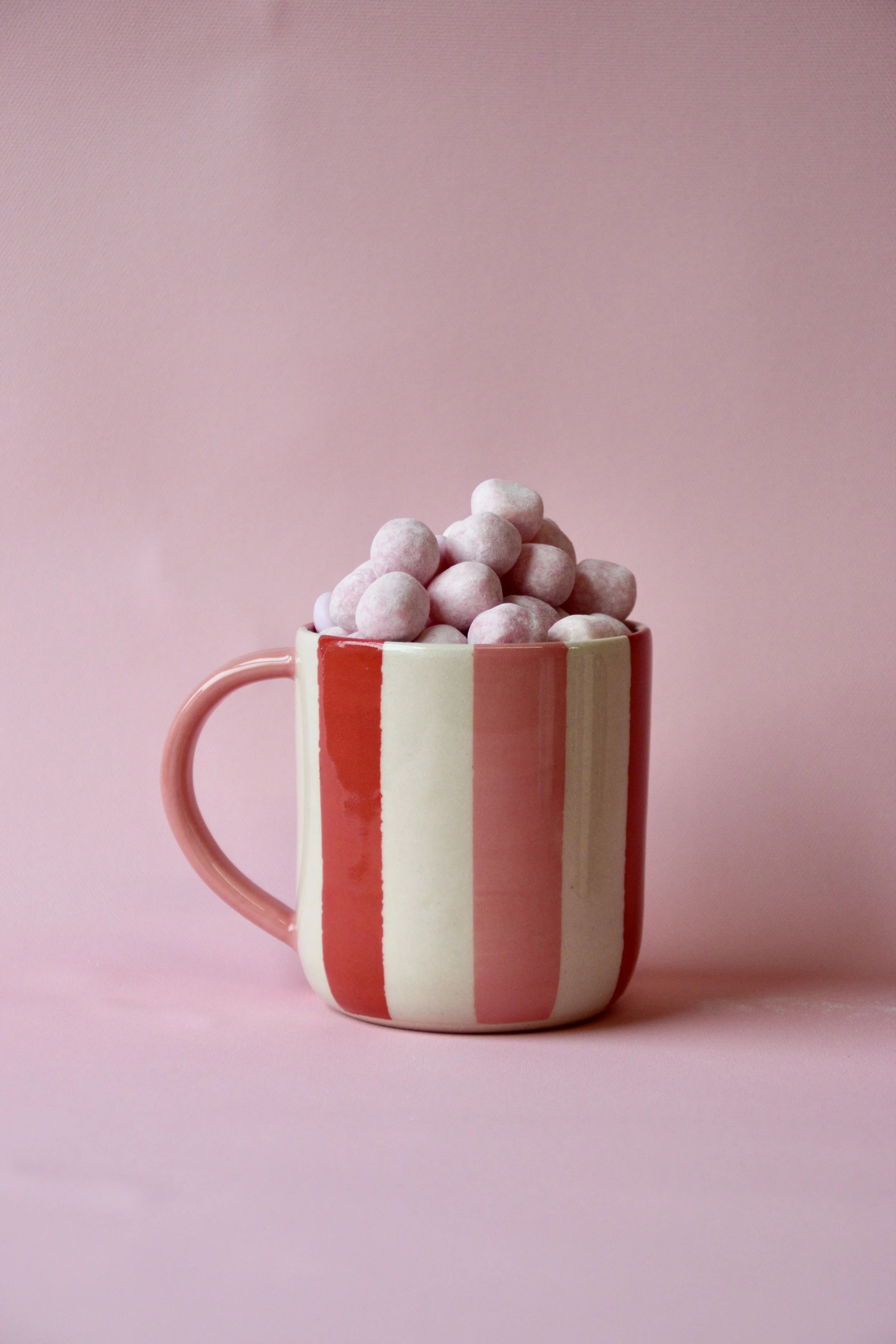 Handmade red and pink striped ceramic mug with matching pink handle, filled with pink sweets, styled on a pink background.