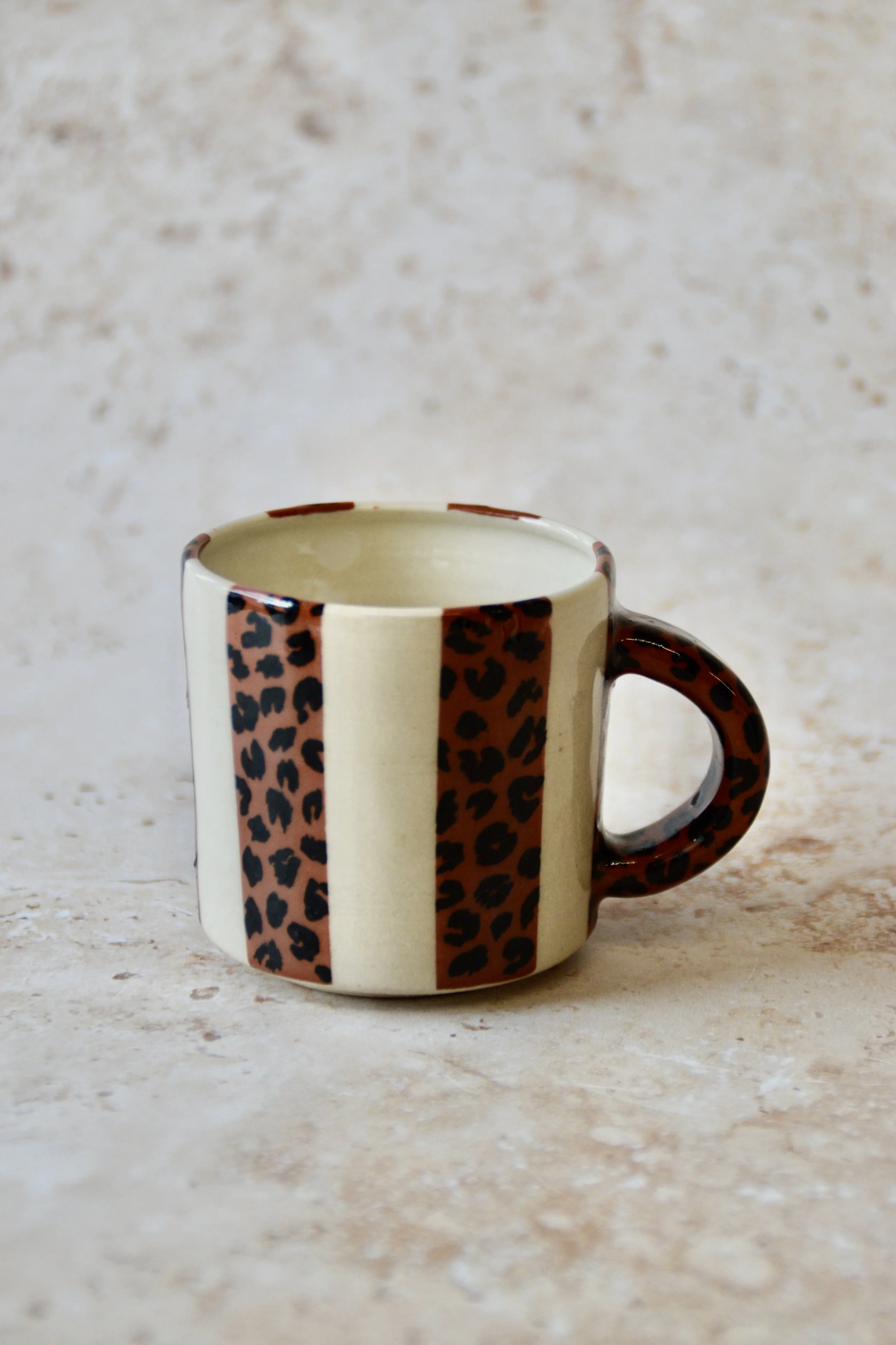 A ceramic mug featuring a cream base with vertical brown leopard print stripes. The handle and rim are brown. This mug is designed for hot drinks and is suitable for use at home or in the office.