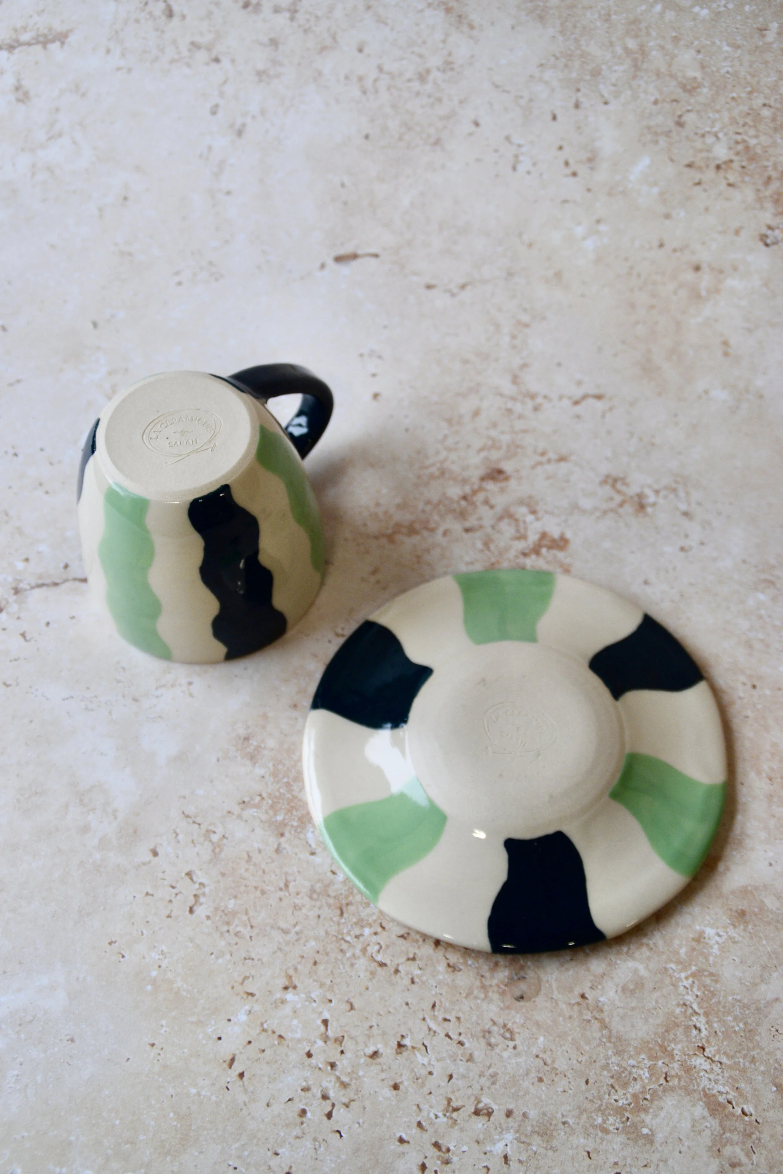 Underside view of a handmade ceramic espresso cup and matching saucer showing the base and maker’s stamp, with wavy green stripe pattern visible on a cream background.