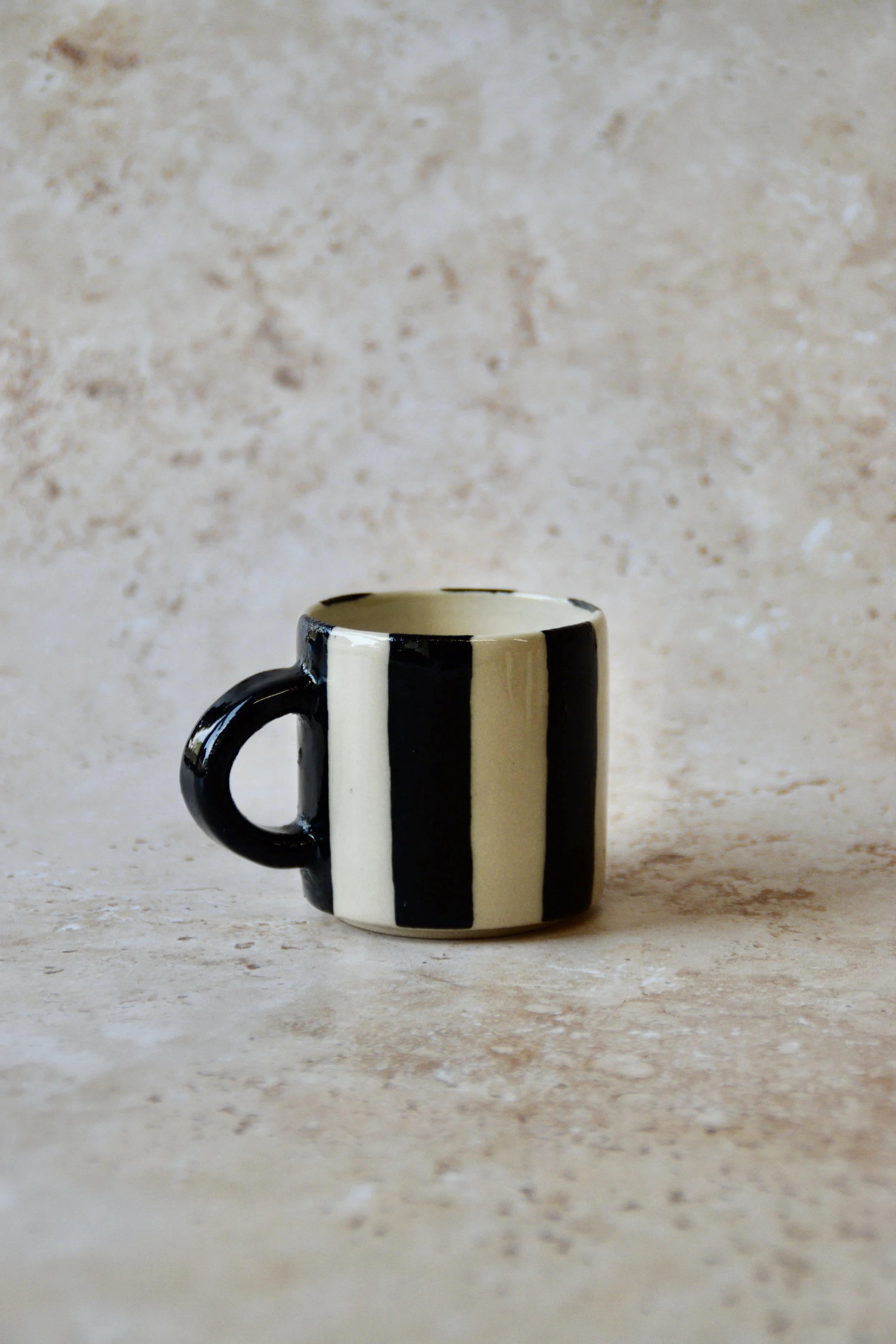 A small ceramic mug with a black handle and vertical black and cream stripes. The mug has a glossy finish and a simple, modern design. The mug is suitable for coffee or tea.