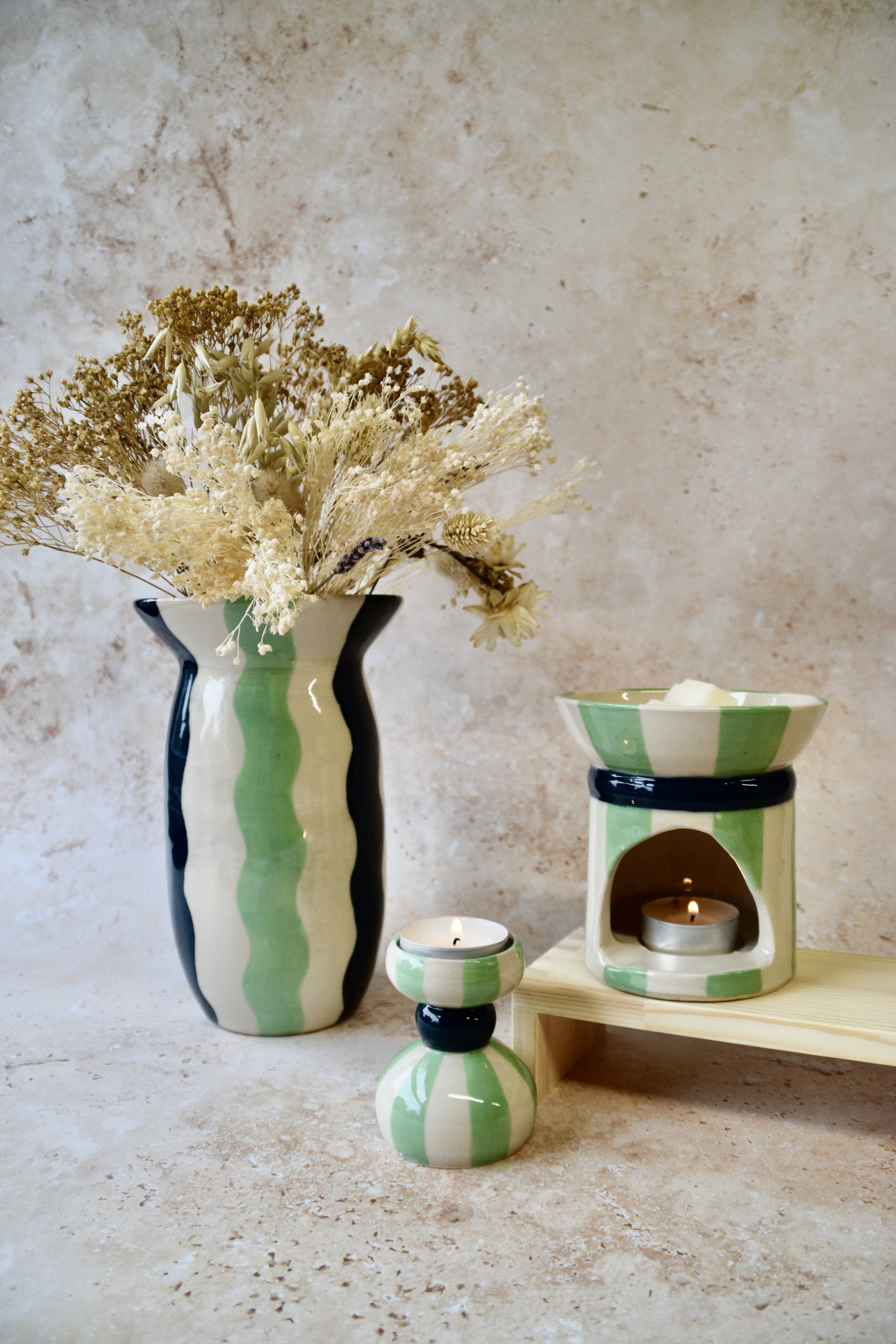 Handmade striped green ceramic collection featuring a vase with dried flowers, a wax burner and a small candlestick on a stone surface.