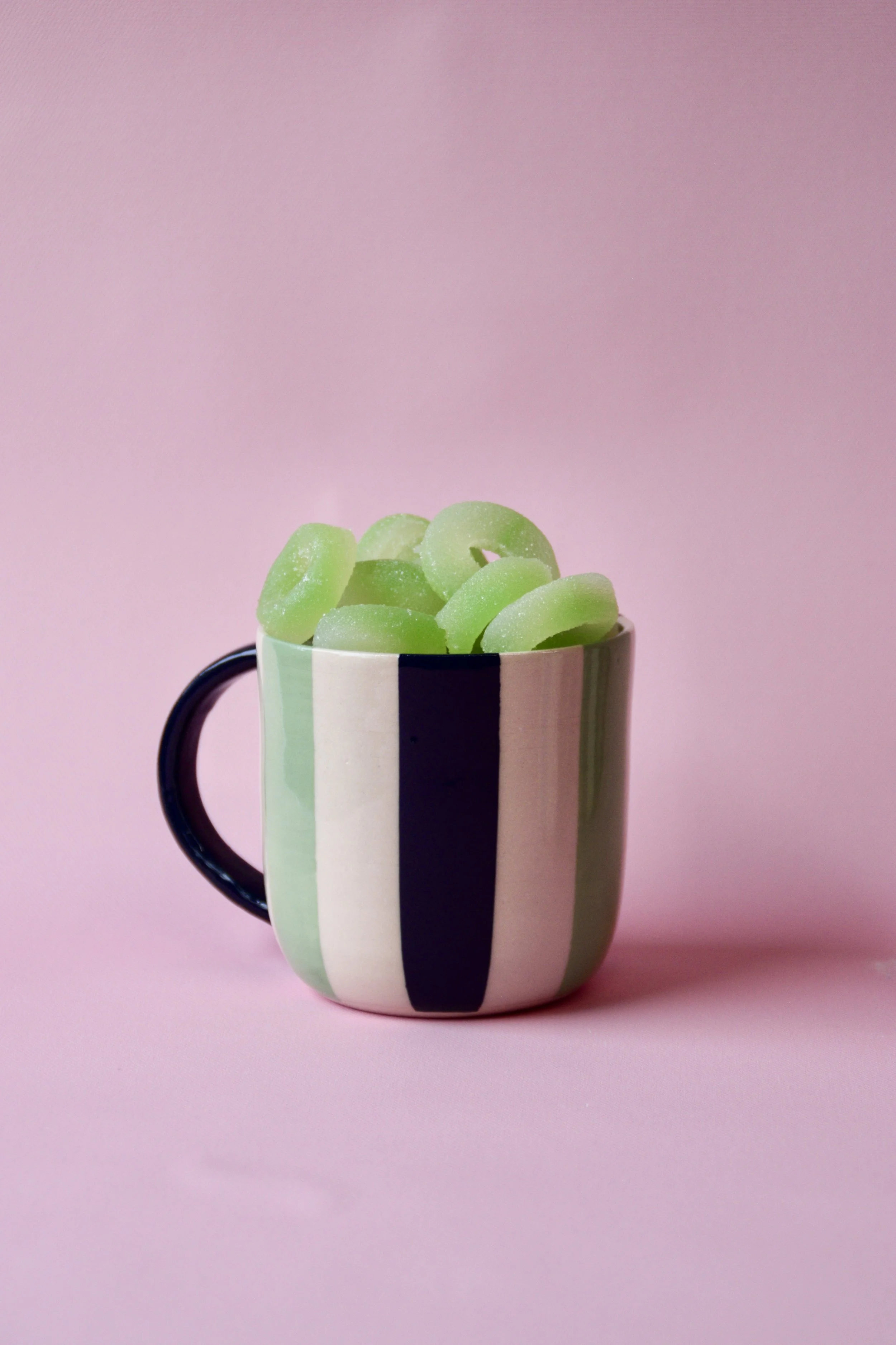 Handmade green and cream striped ceramic mug with dark green handle, filled with green gummy sweets, styled on a pink background.