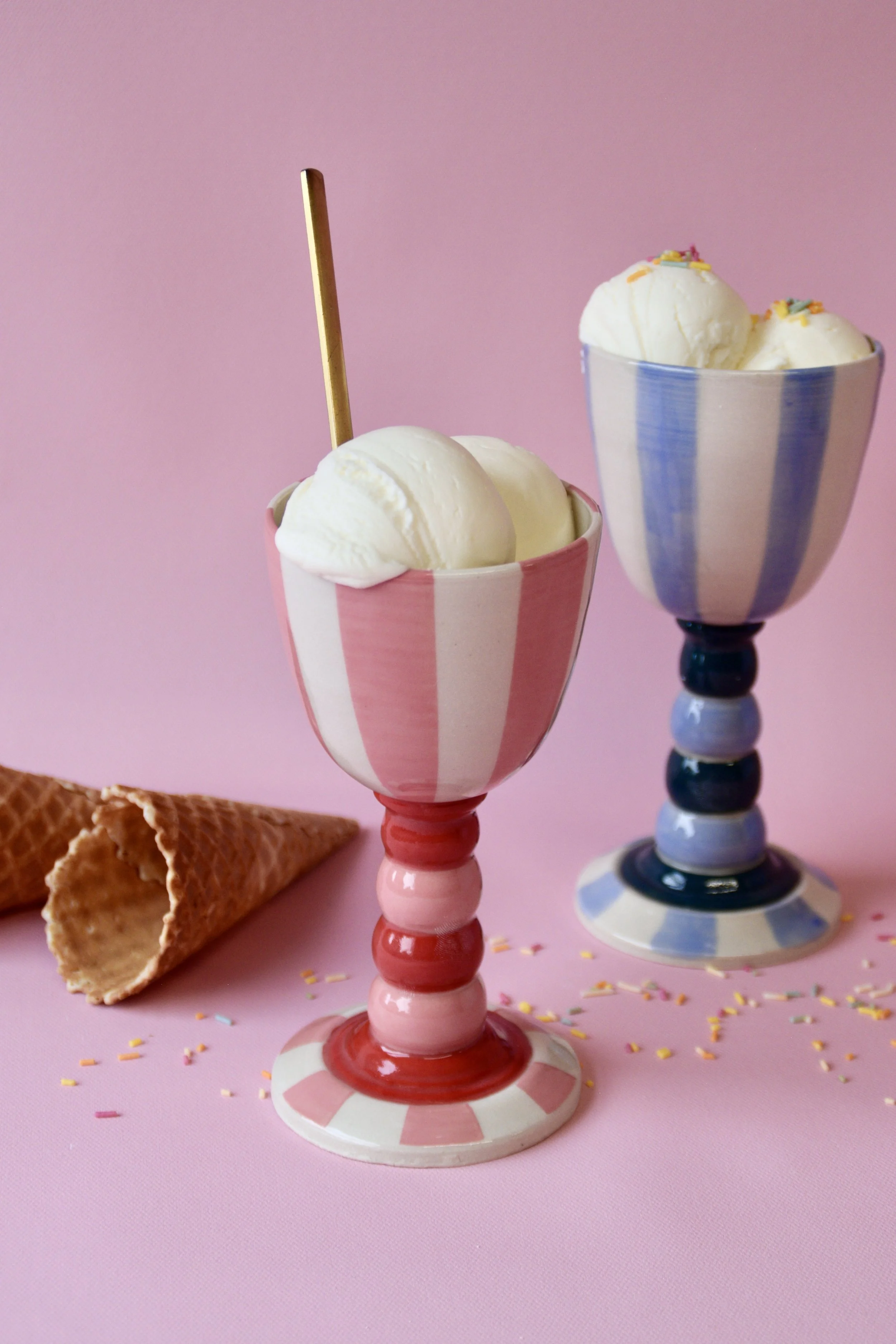Two handmade striped ceramic Carnival goblets filled with whipped cream, photographed on a pink background with an ice cream cone.