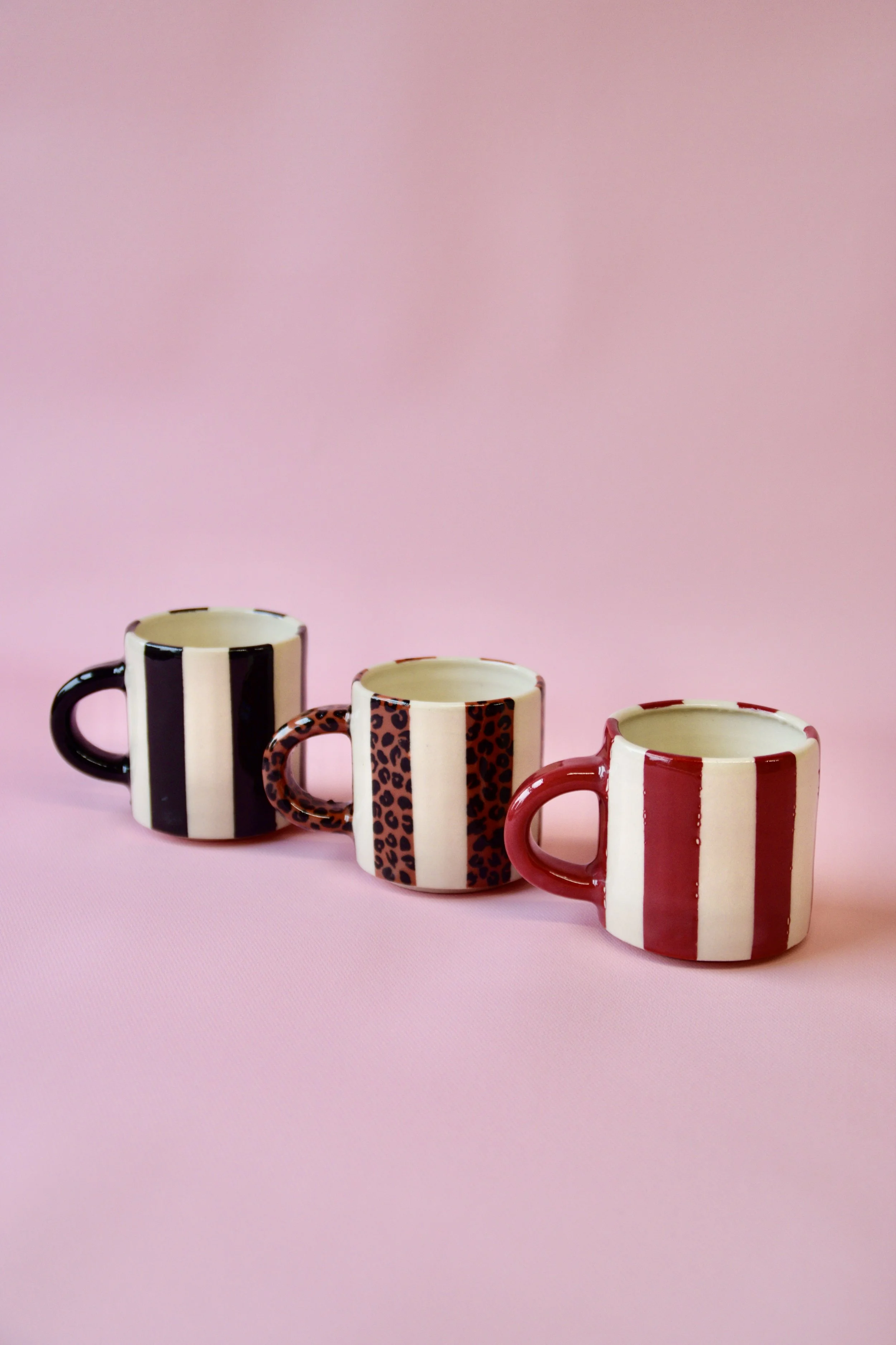 Three handmade ceramic espresso cups with bold striped and leopard pattern designs, displayed on soft pink background.