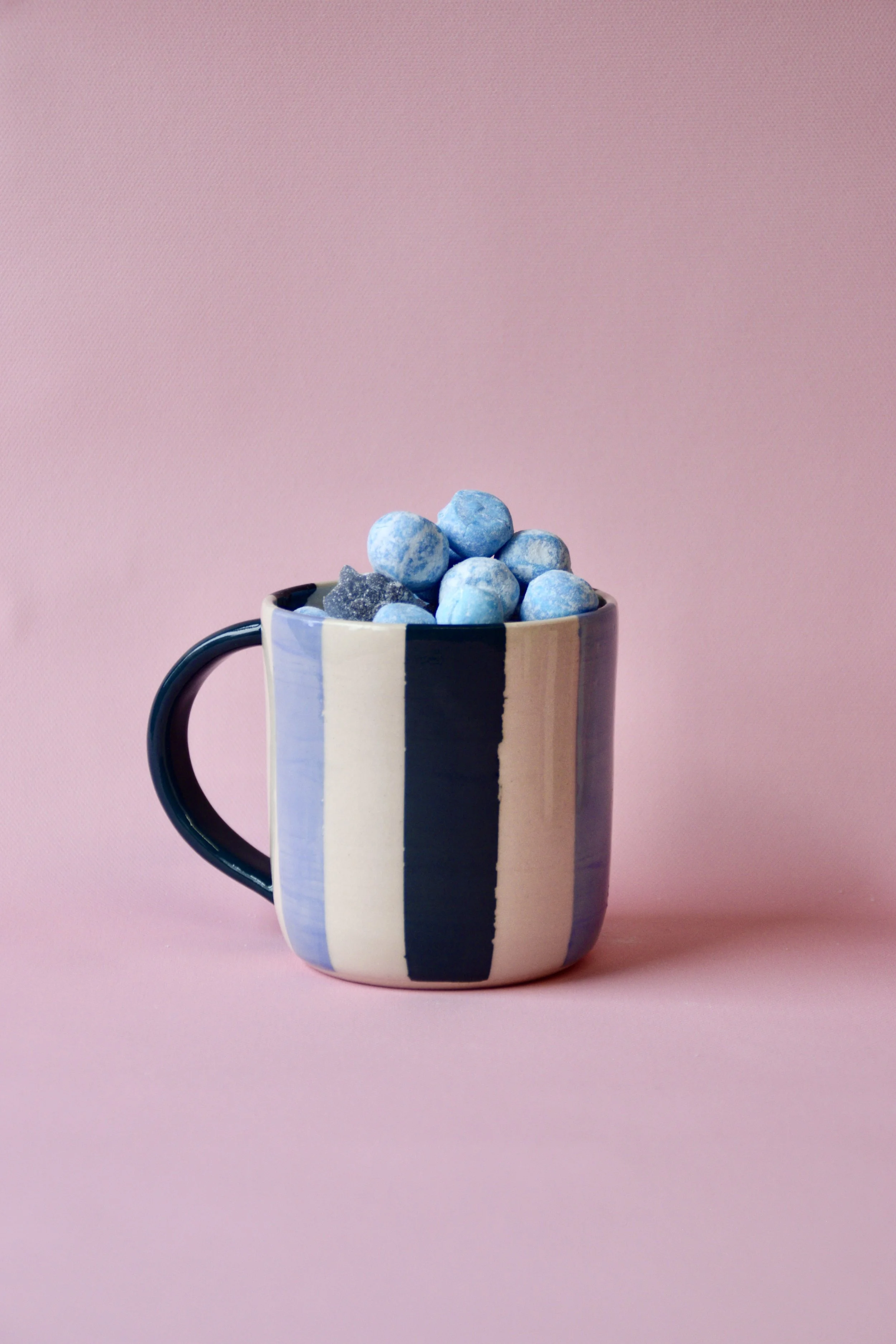 Handmade blue and cream striped ceramic mug with dark teal handle, filled with blue sweets, styled on a pink background.