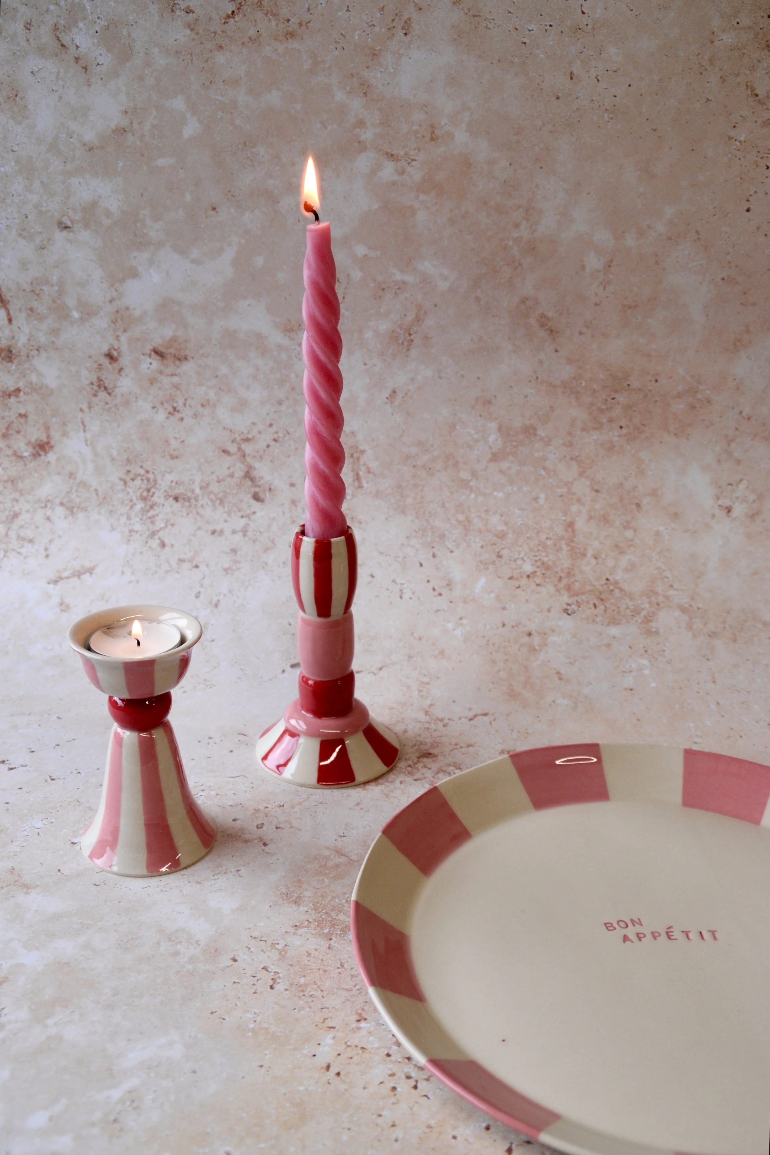 Handmade striped ceramic candlestick holder and matching tea light holder beside a red and cream striped dinner plate on a stone background.