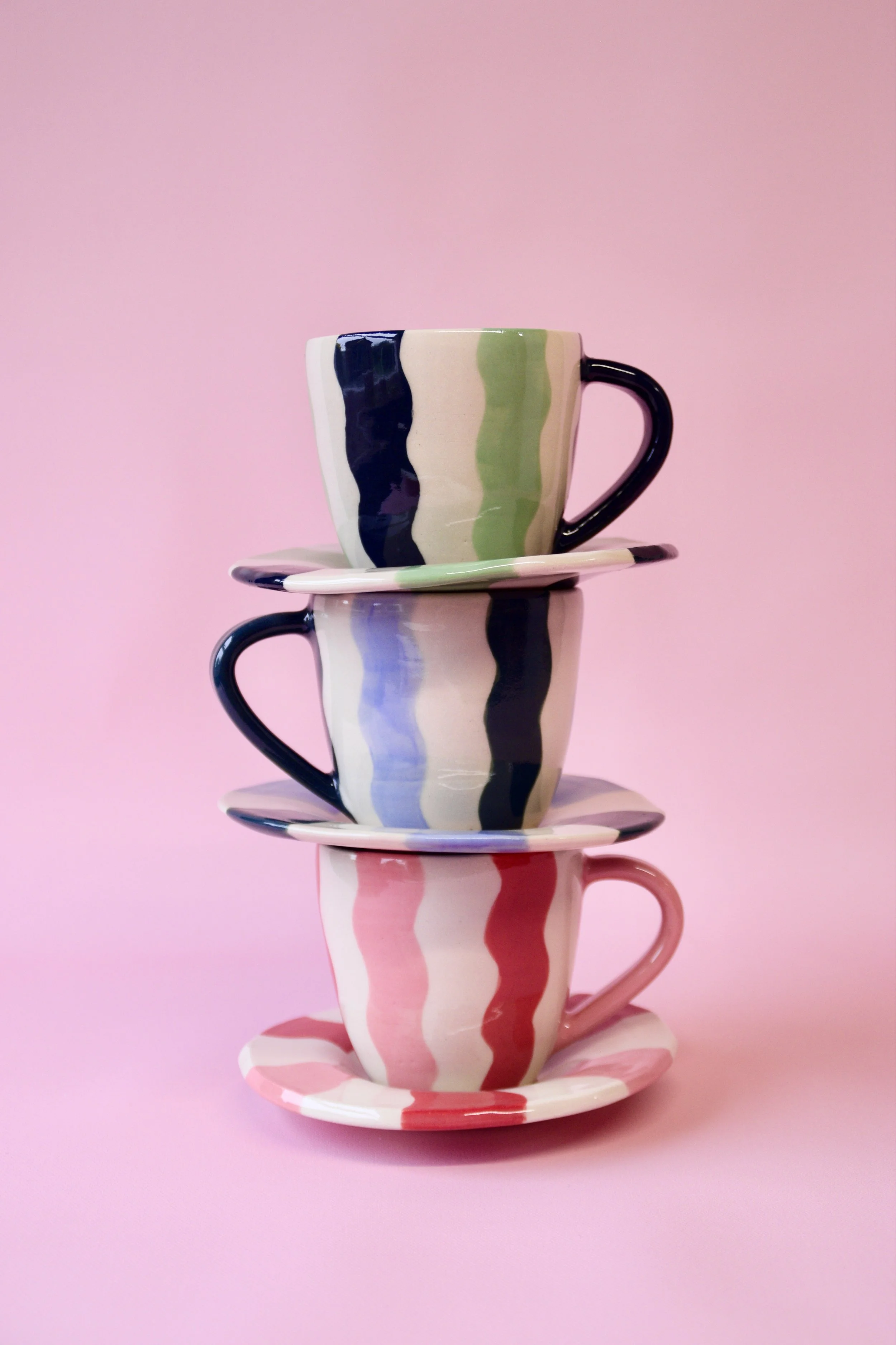 Three handmade striped ceramic cups stacked with matching saucers in green, blue and red colourways on a soft pink background.