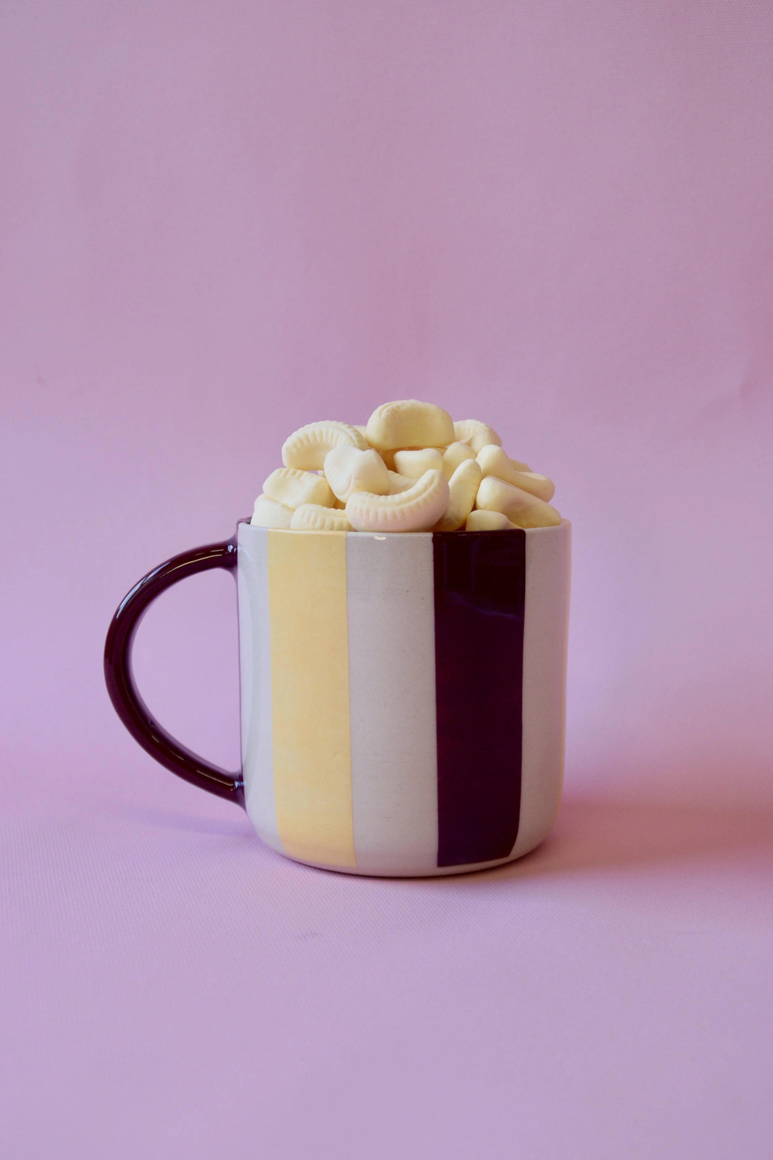 Handmade striped ceramic mug with vertical burgundy and butter yellow stripes on a creamy white background, filled with mini bananas and photographed against a soft pink backdrop.