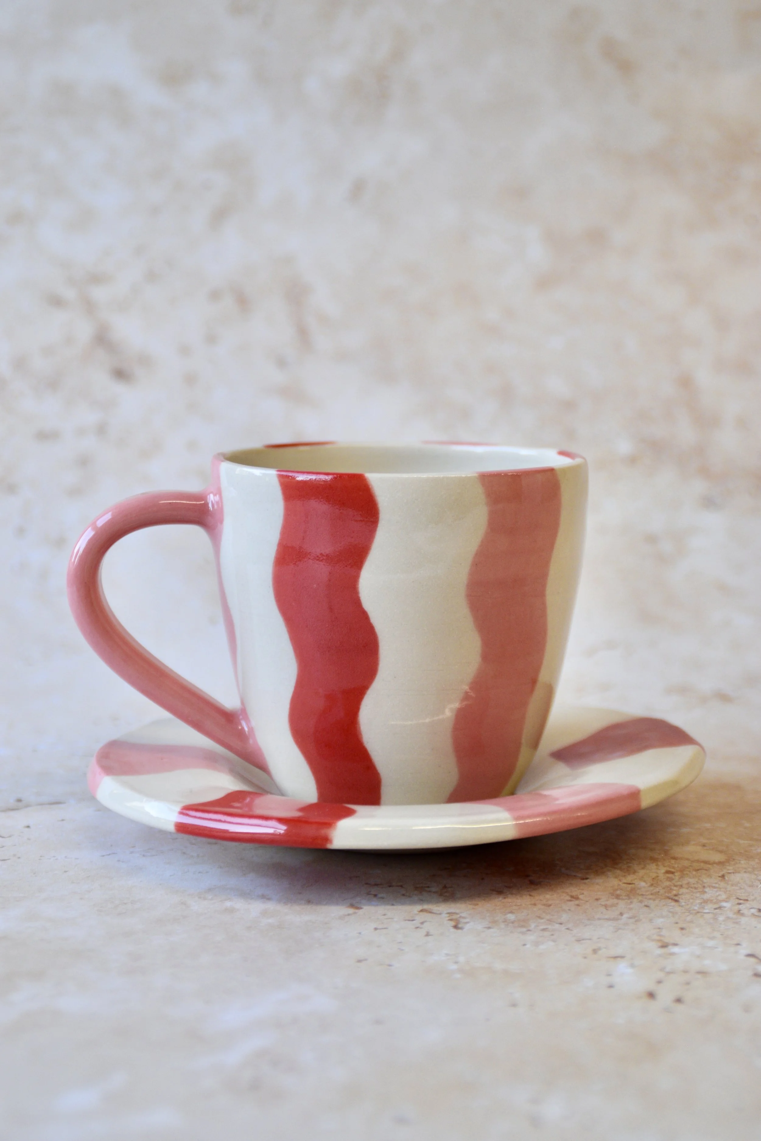Handmade wavy striped ceramic coffee cup and saucer on a neutral stone background.