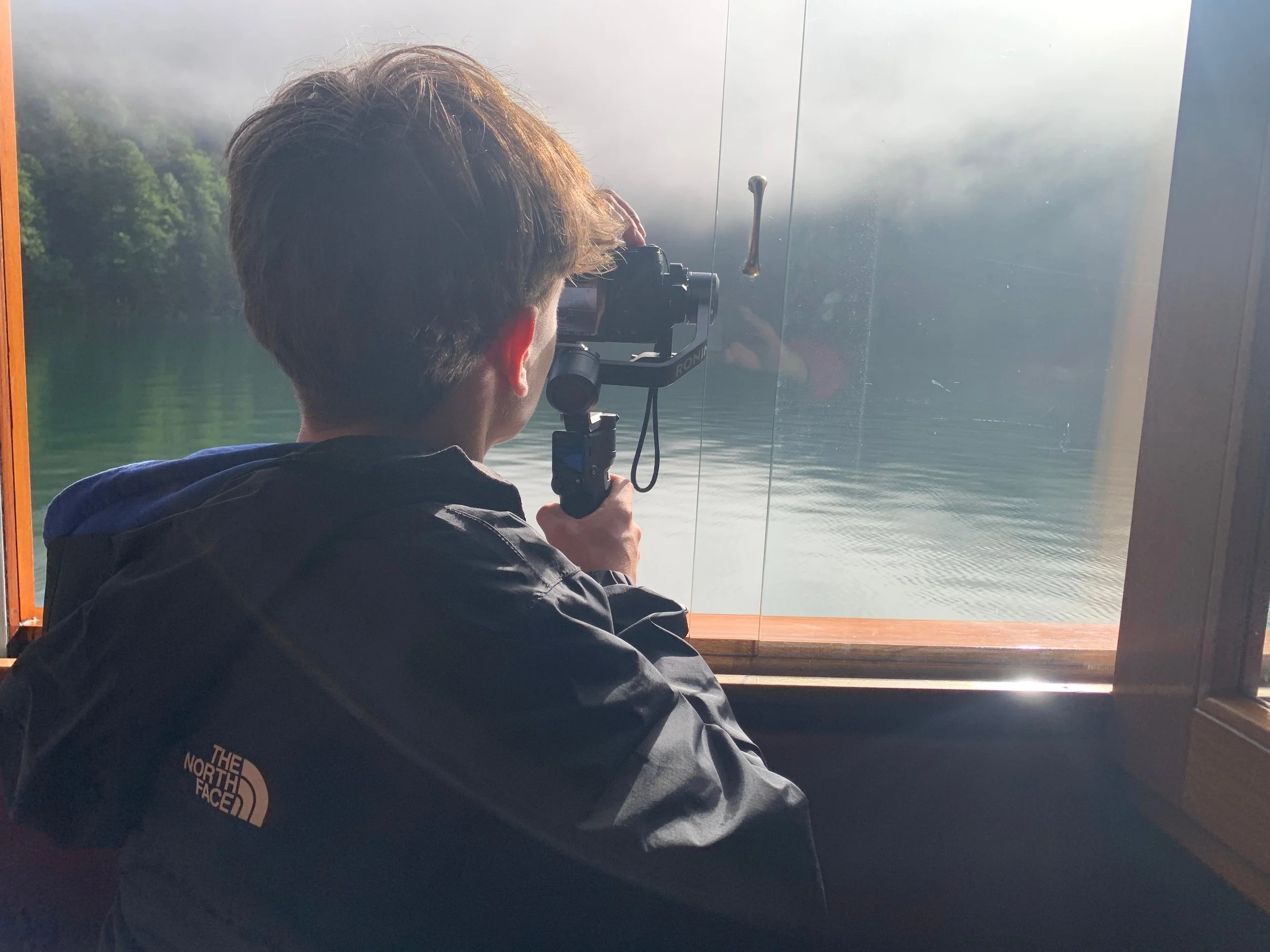 Junge Person filmt mit Kamera durch ein Fenster auf einen ruhigen See mit umliegendem grünen Wald, bei Nebel. Er trägt eine schwarze Jacke mit North Face Logo.