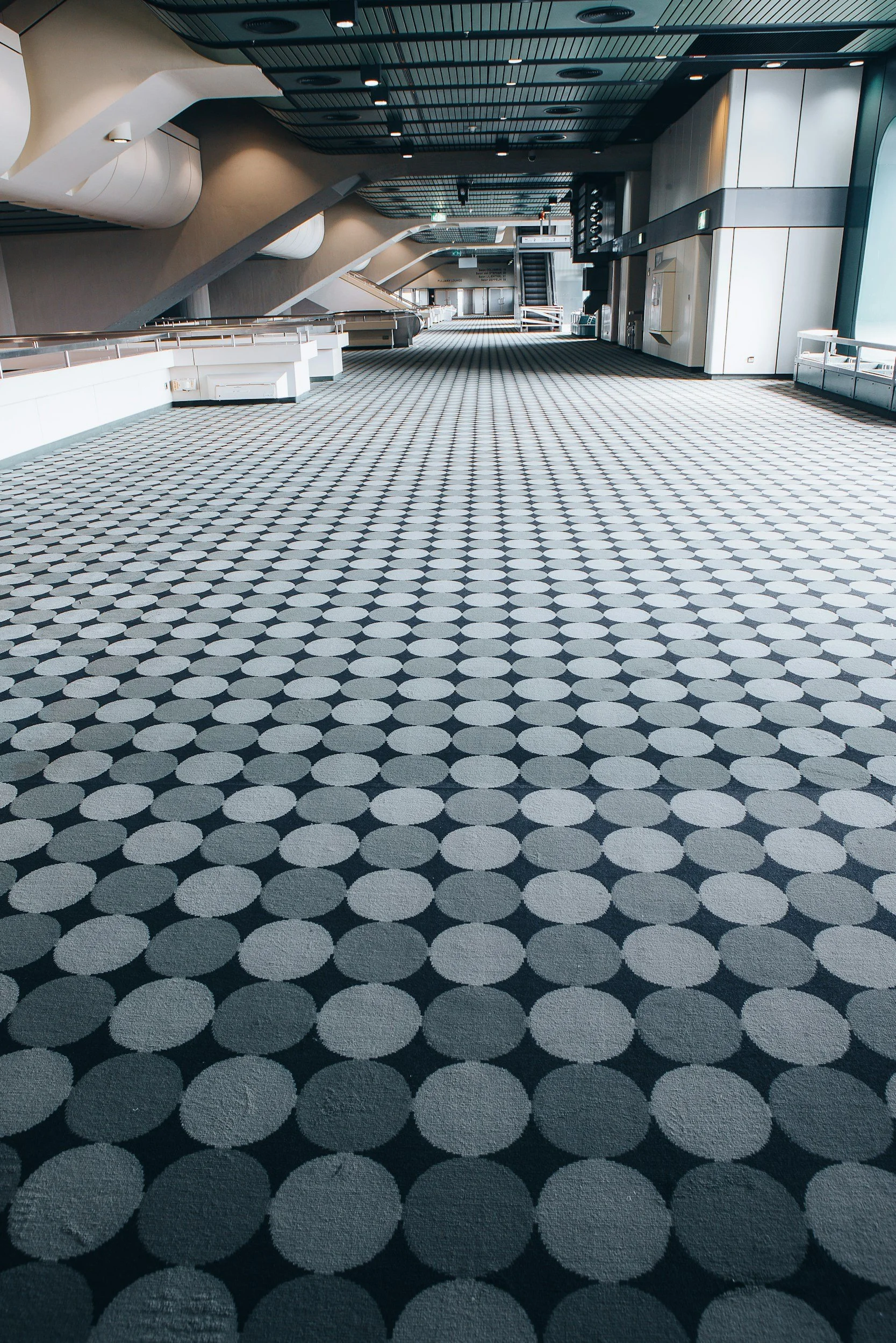Empty modern airport terminal with black and white patterned floor, escalators, and large windows.