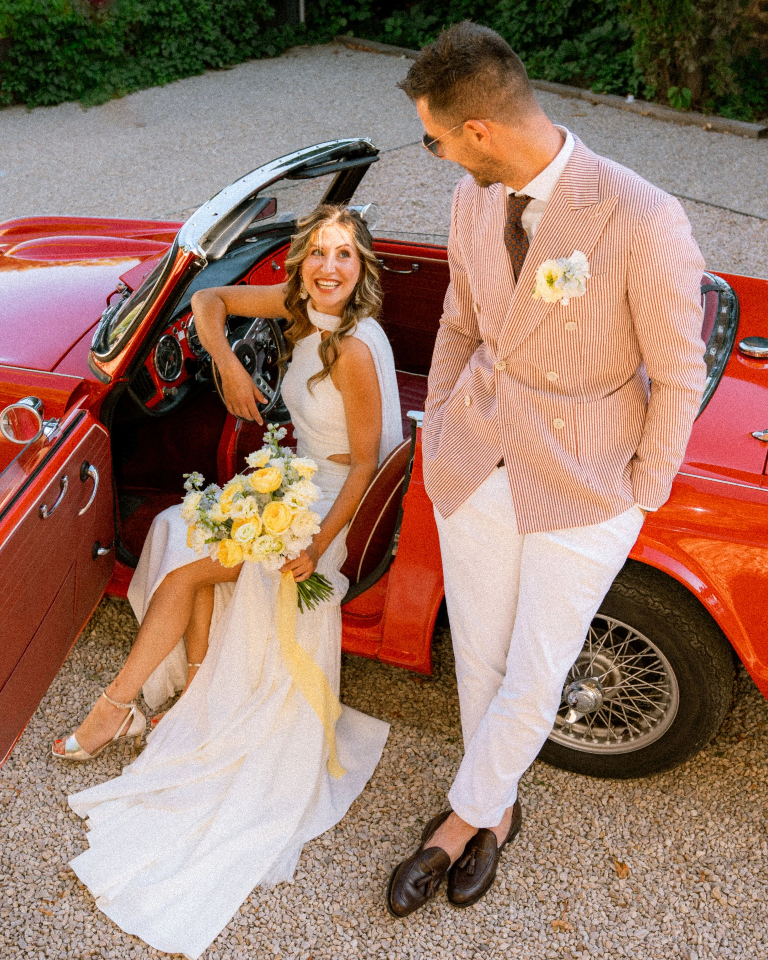 Jeune couple de mariés avec la femme assise dans une voiture rouge classique, tenant un bouquet de fleurs jaunes, et l'homme debout à côté, portant un costume à rayures, tous deux souriants, en extérieur