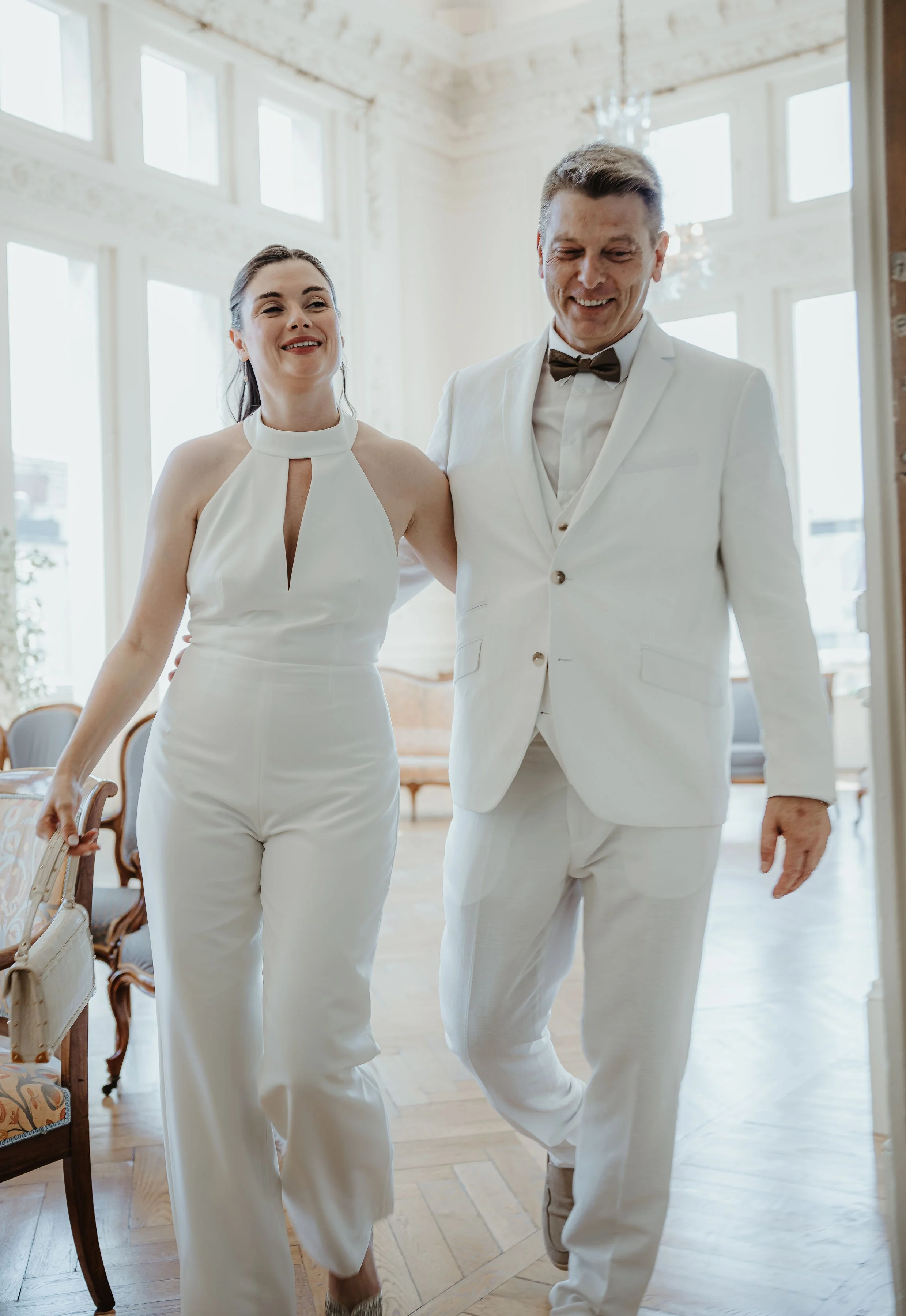 Un couple en tenue de mariage marchant dans une salle lumineuse avec de grandes fenêtres et des meubles anciens.