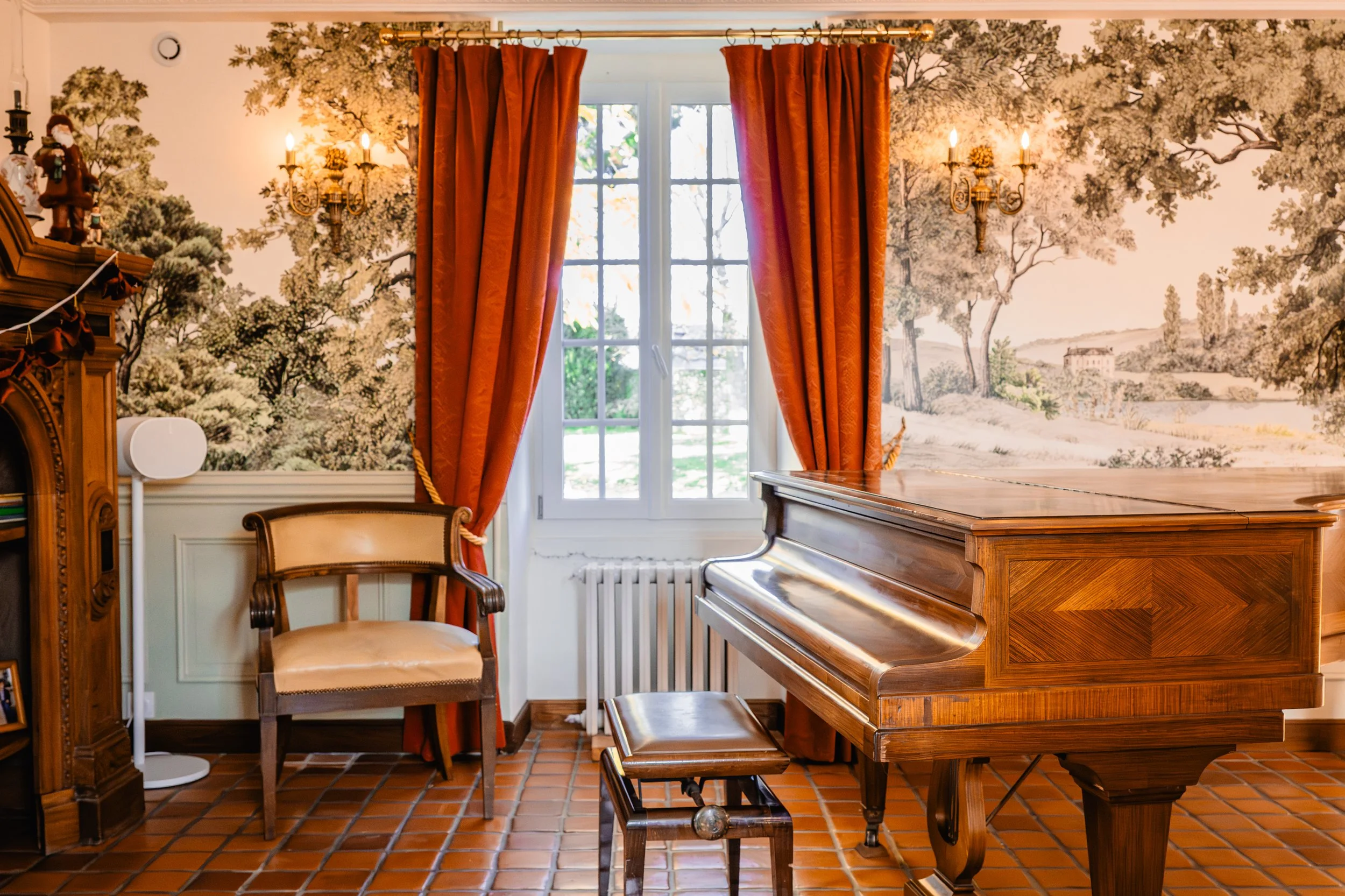 Salle de musique avec un piano en bois, une chaise en osier avec un coussin, des rideaux rouges, une tapisserie murale représentant une scène champêtre, et des appliques murales éclairant la pièce.