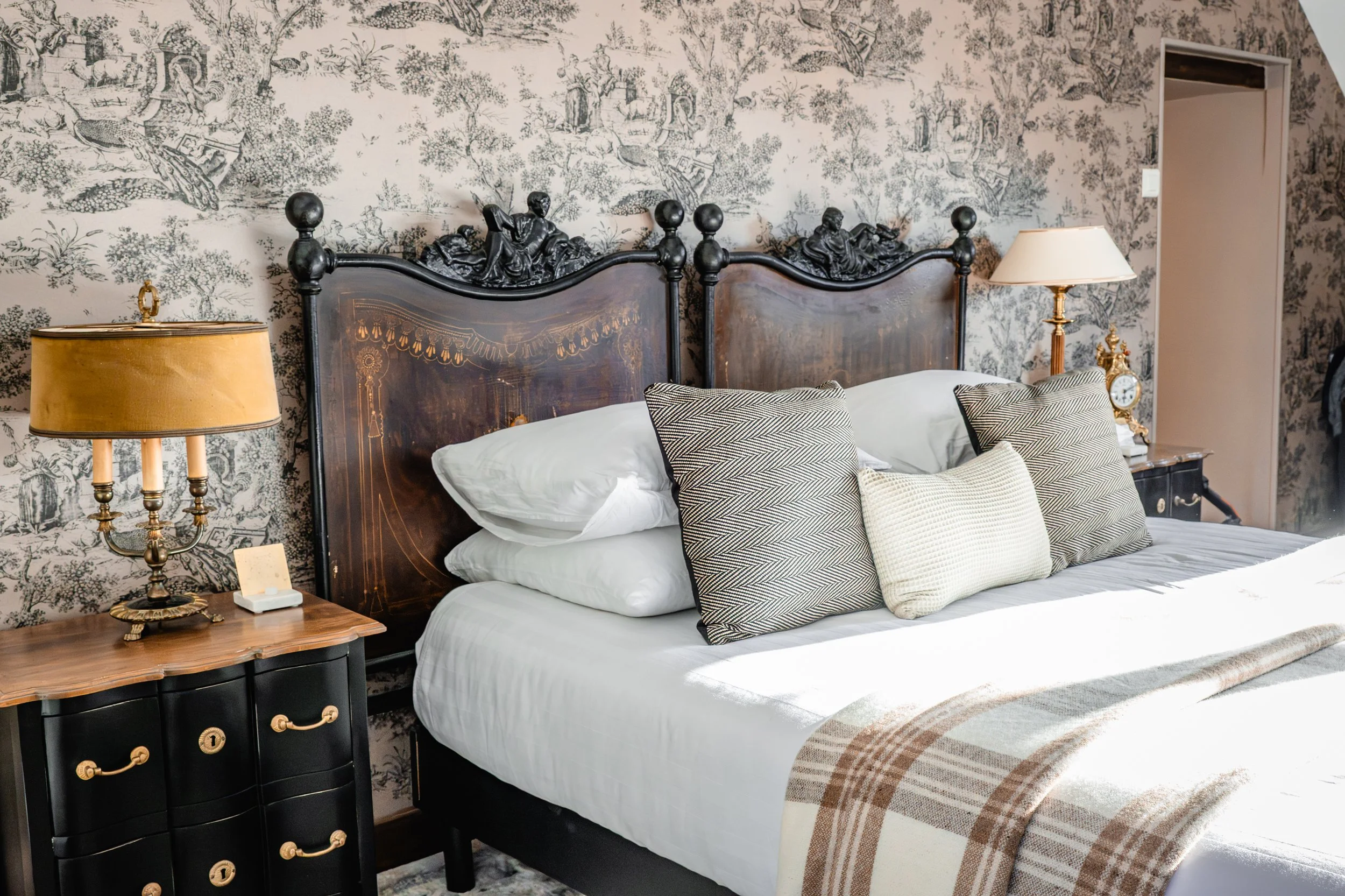 Une chambre élégante avec un lit à grand headboard en bois, plusieurs oreillers, deux lampes de chevet avec abat-jour, et un papier peint aux motifs de paysage en noir et blanc.