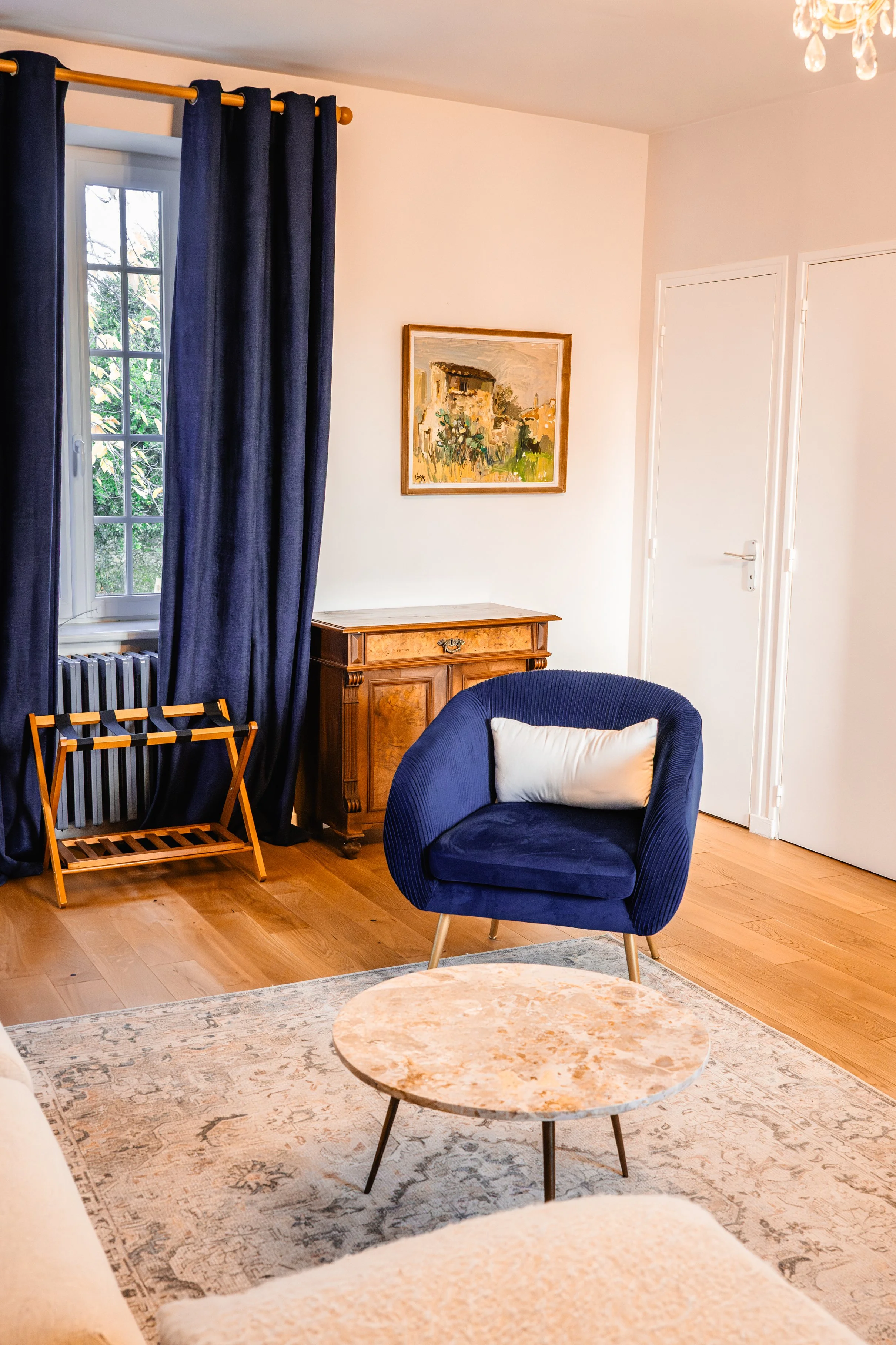 Salon avec un fauteuil bleu, un tapis beige, une table en marbre, une fenêtre avec rideaux bleus, une petite table en bois, un tableau avec une peinture de style impressionniste et un lustre au plafond.