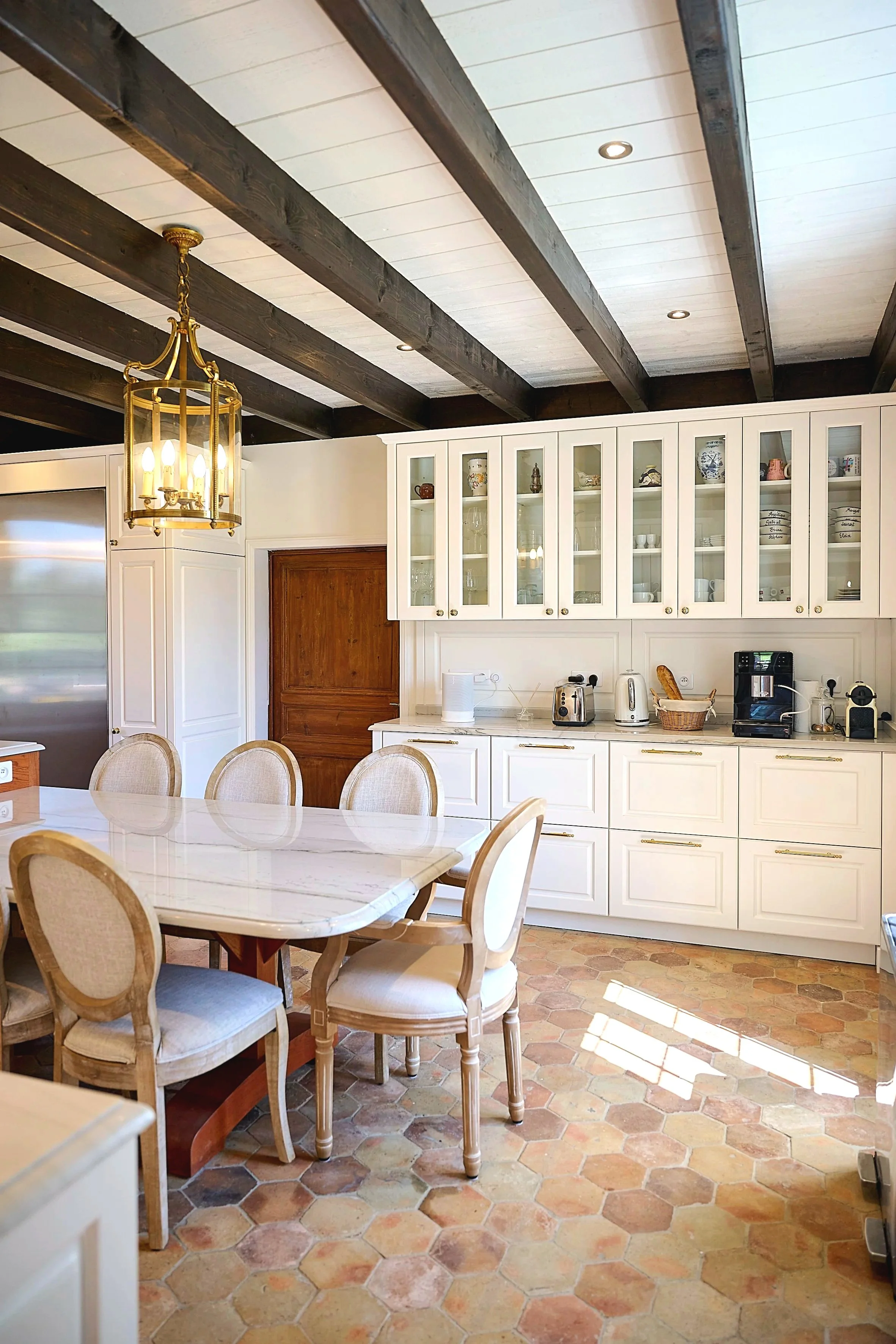 Cuisine lumineuse avec table en marbre, chaises en bois beige, armoires blanches avec portes vitrées, plafond avec poutres en bois foncé, lustre en métal doré, appareils électroménagers sur le comptoir.