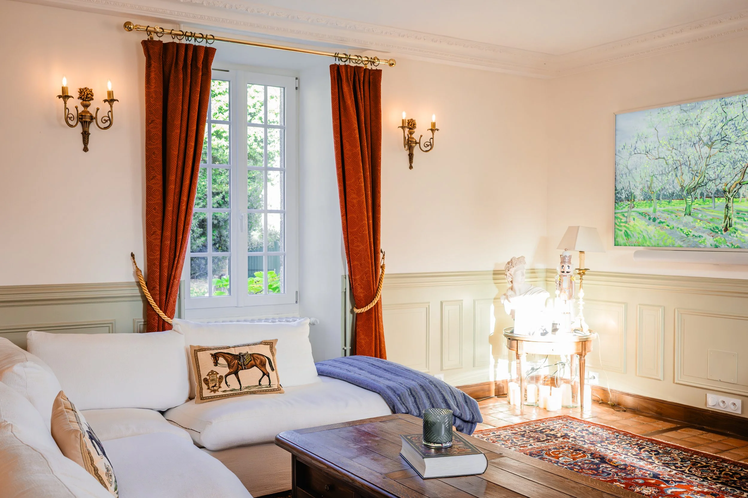 Salon lumineux avec canapé blanc, coussin à motif cheval, table en bois avec livre et bougie, fenêtre avec rideaux rouges, tableau de paysage verdoyant, lampe et sculpture sur une table
