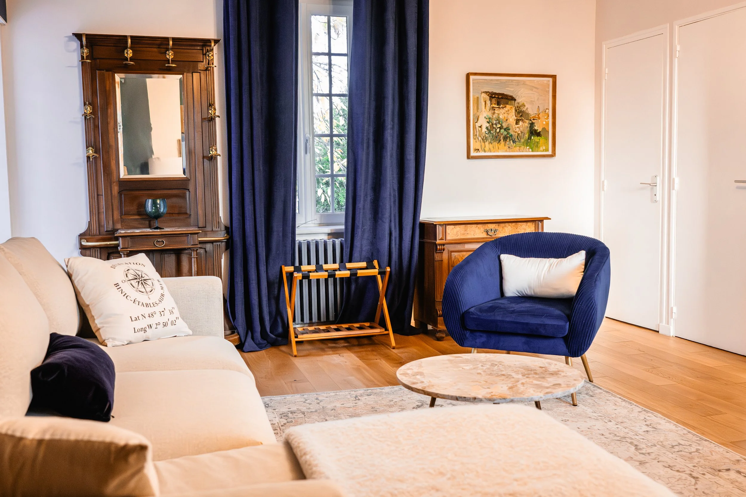 Salon avec canapé beige, fauteuil bleu, tables en marbre, rideaux bleus, peinture sur le mur, meubles en bois et fenêtre avec vue sur des arbres.