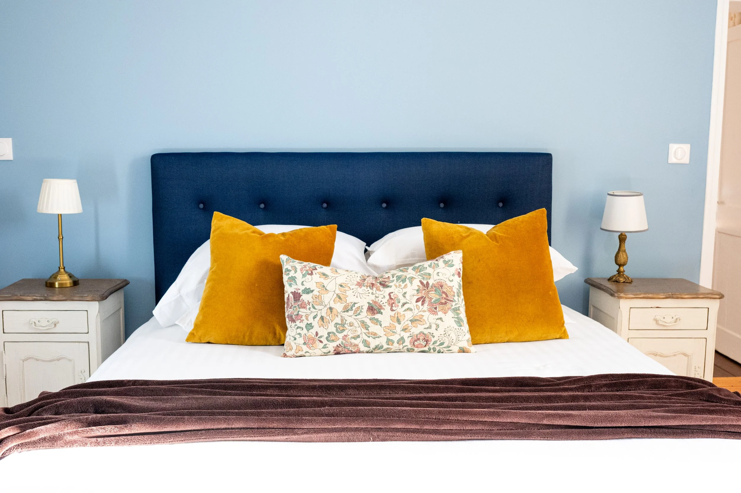 Lit double avec un tête de lit bleue, deux lampes de chevet, deux coussins orange et un coussin floral, couvre-lit marron.