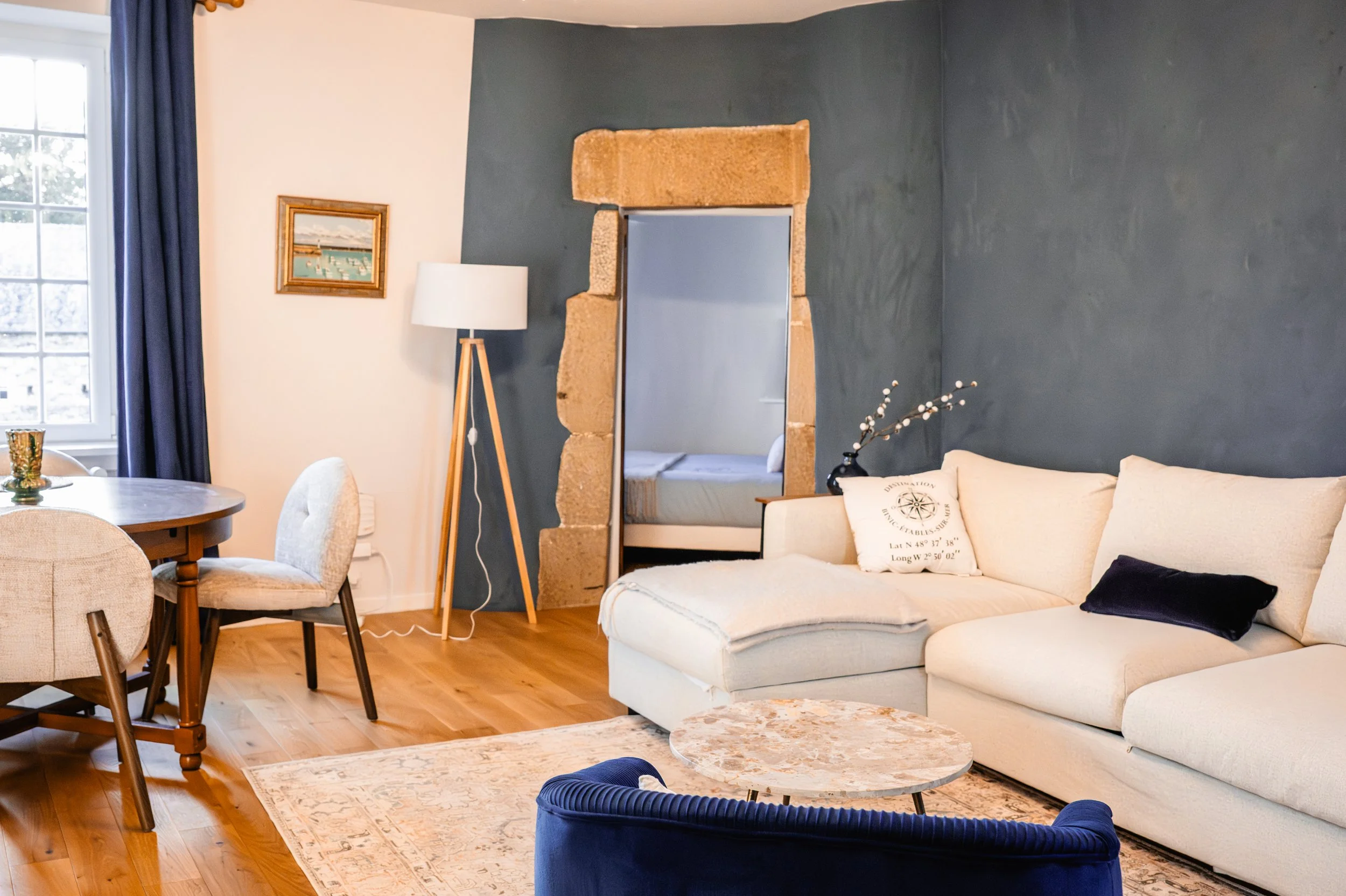 Salon lumineux avec un canapé blanc en forme de L, table basse en marbre, fauteuil bleu, lampe sur pied à gauche, mur bleu foncé, porte en pierre menant à une chambre, table ronde en bois avec un vase aux fleurs, cadre au mur à gauche, tapis beige, fenêtre avec rideaux bleus.