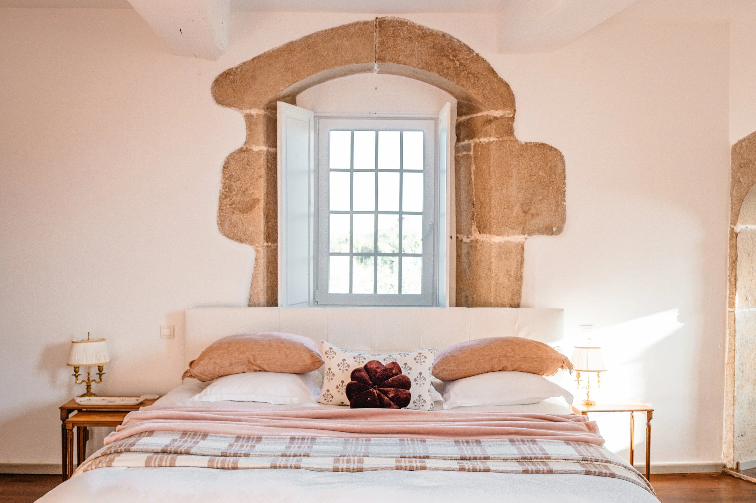 Chambre à coucher avec lit, oreillers et table de nuit, fenêtre avec volets, murs en pierre et plafond voûté.
