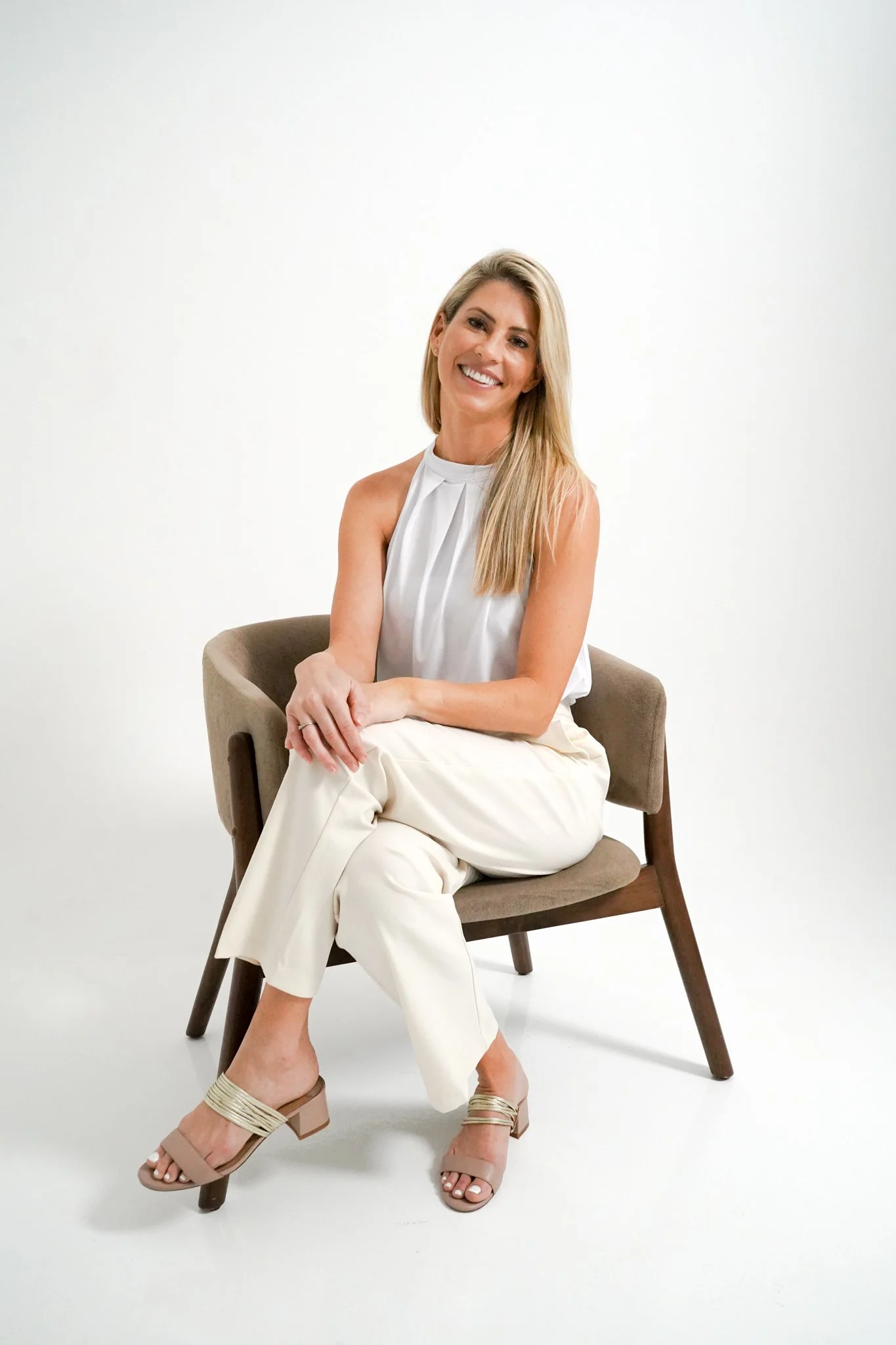 A woman with long blonde hair sitting on a brown chair, wearing a white sleeveless top and cream-colored pants, smiling with her legs crossed.