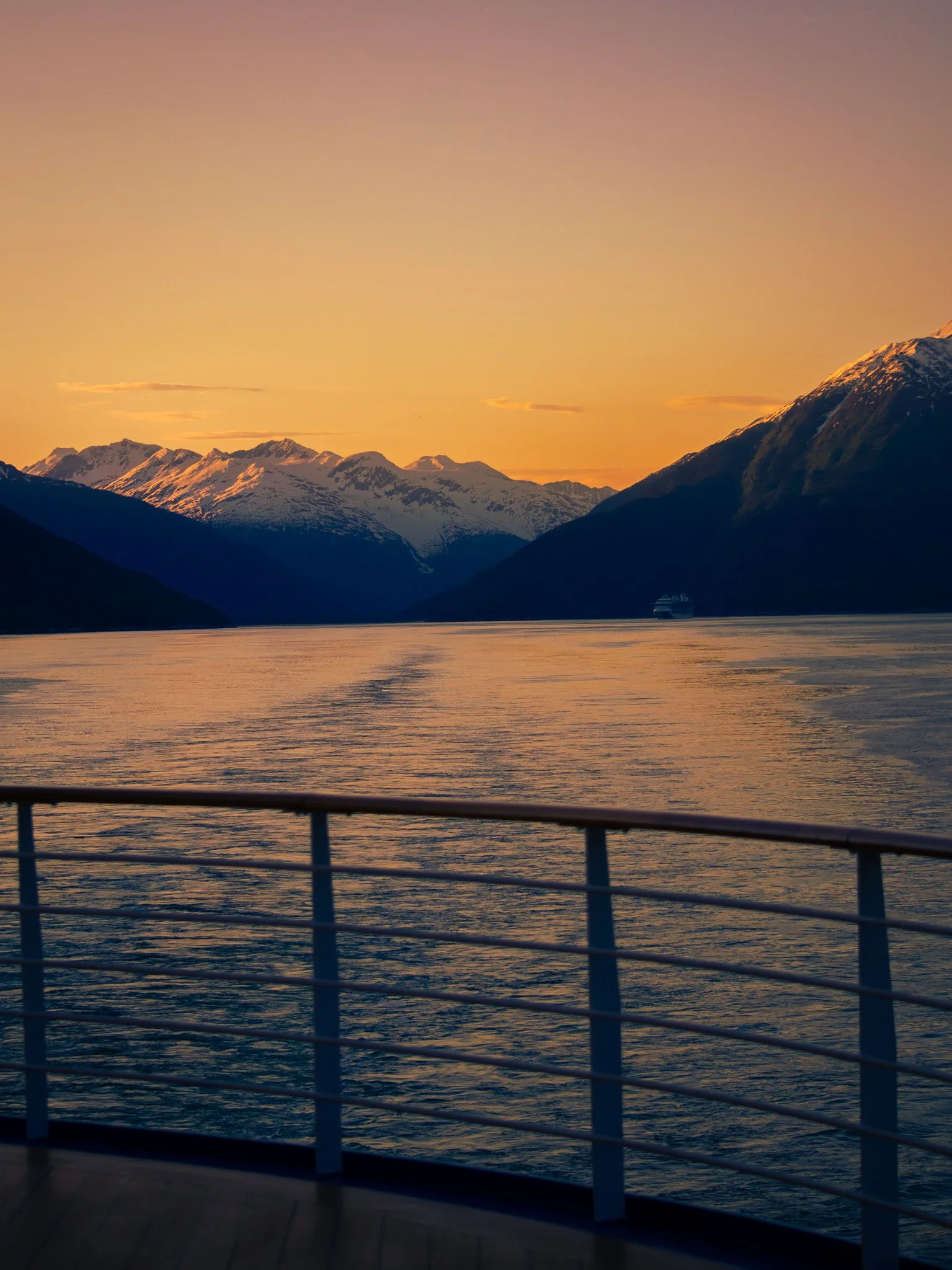 Sunset over a fjord with snow-capped mountains in the background and a ship in the distance.