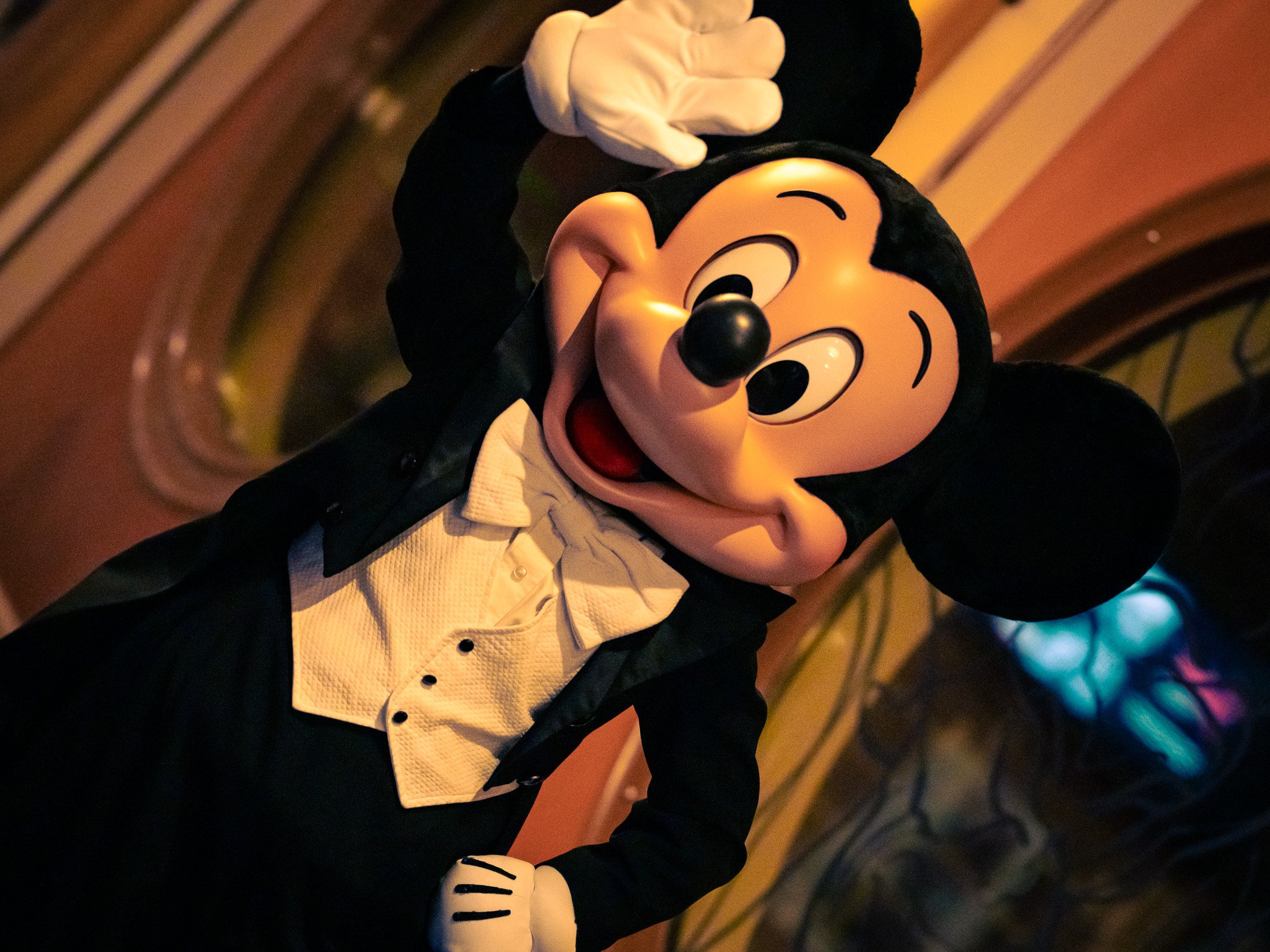 Mickey Mouse wearing a tuxedo with white shirt and black bowtie, waving and smiling.