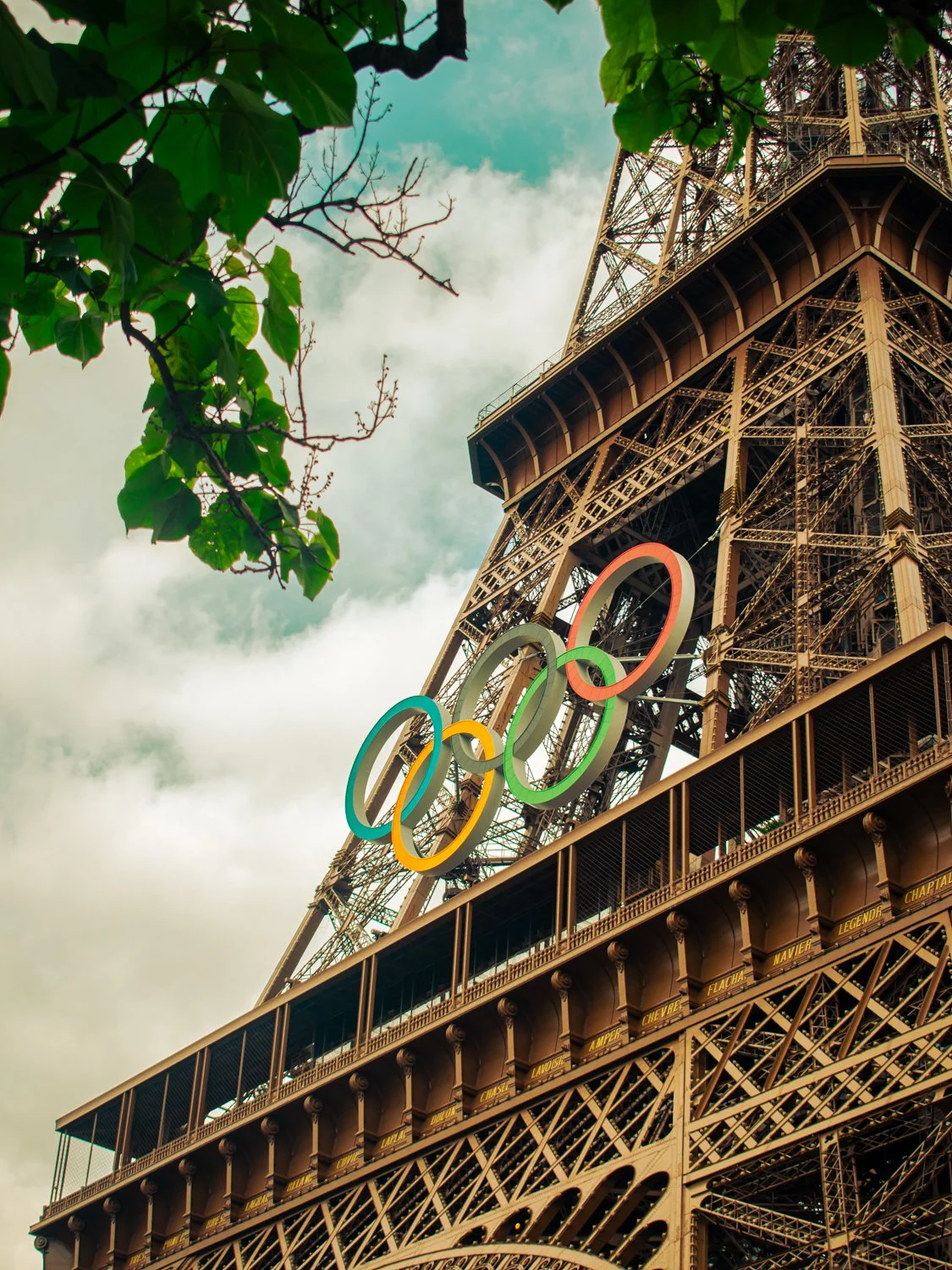 Eiffel Tower with Olympic rings themed decorations in Paris, France, surrounded by green leaves and cloudy sky.