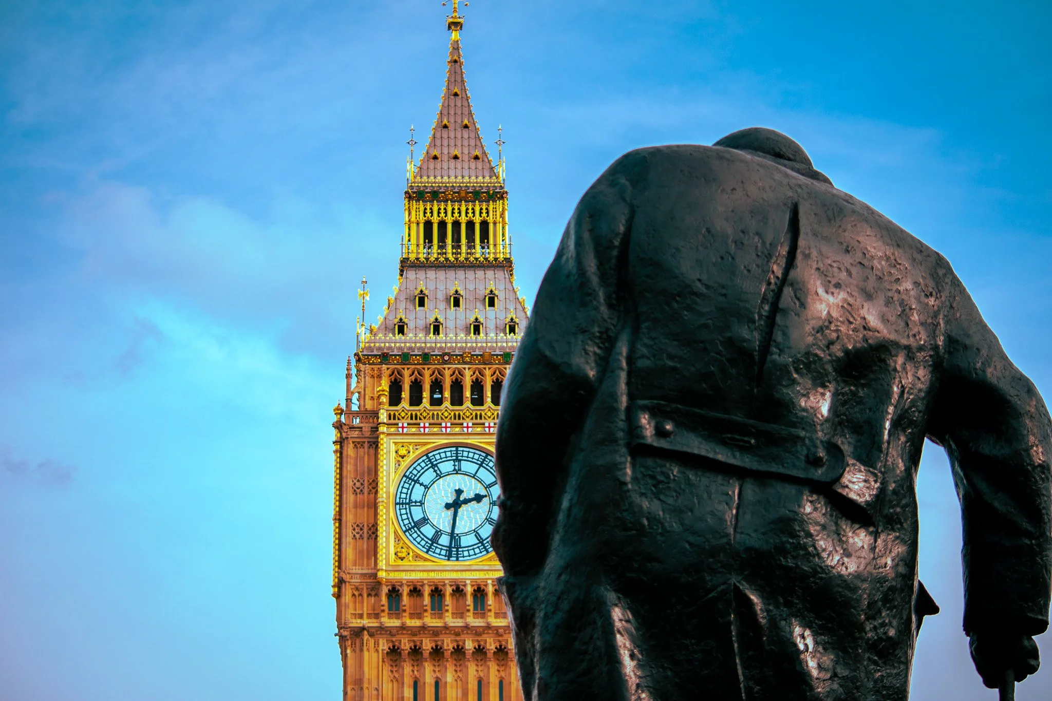 A bronze statue of Winston Churchill in a coat with his back turned, facing the Big Ben clock tower in London, England, against a blue sky.