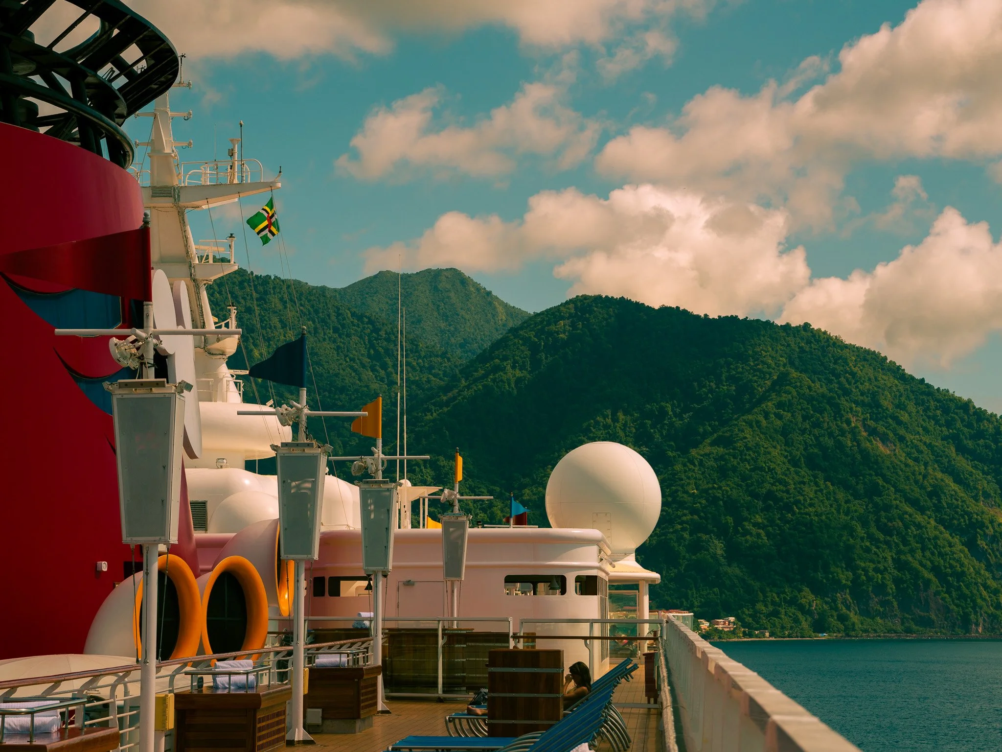 View of a cruise ship deck with seating and antenna equipment, with green mountain landscape in the background and a partly cloudy sky. Captured on the Disney Magic ship in Dominica.