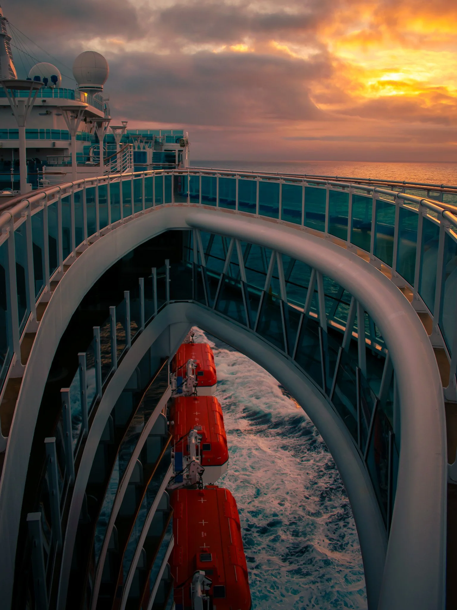 A view of the deck and lifeboats of a cruise ship during sunset, with the ocean and sky in the background.