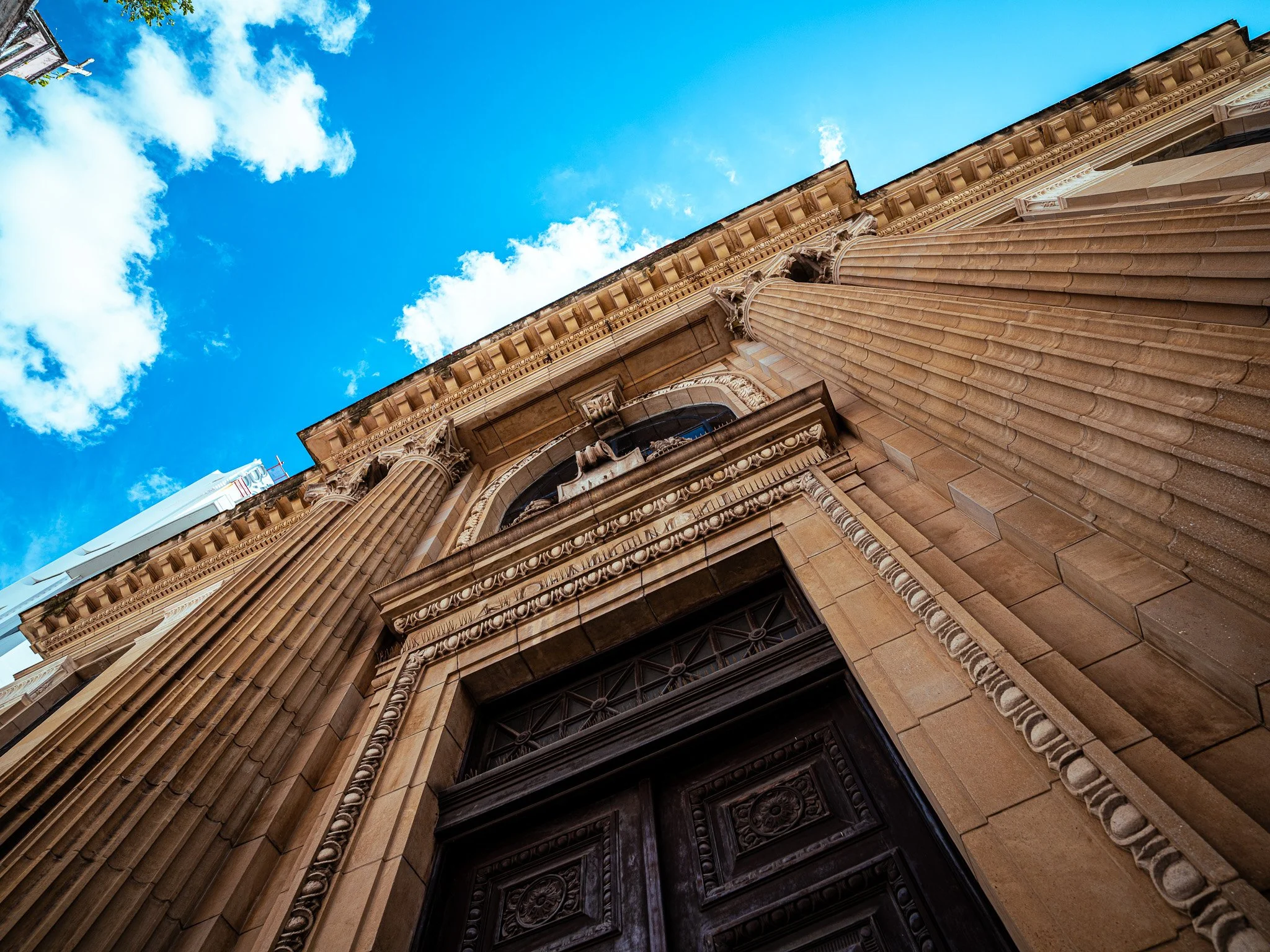 Low angle view of an ornate building facade with large columns, decorative stonework, and a dark wooden door, against a bright blue sky with white clouds.