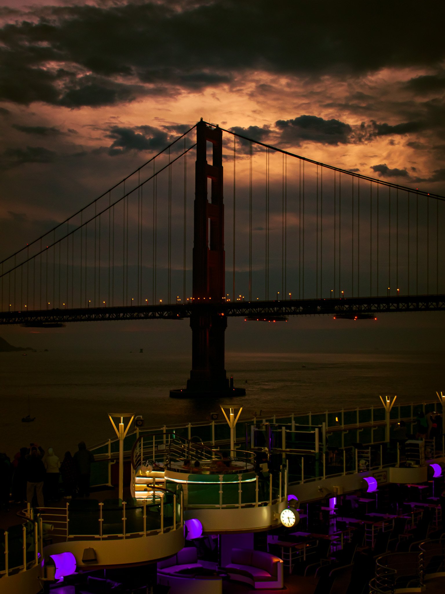 Sunset view of the Golden Gate Bridge in San Francisco with a cloudy sky in the background, seen from a cruise ship deck illuminated with purple and yellow lights.
