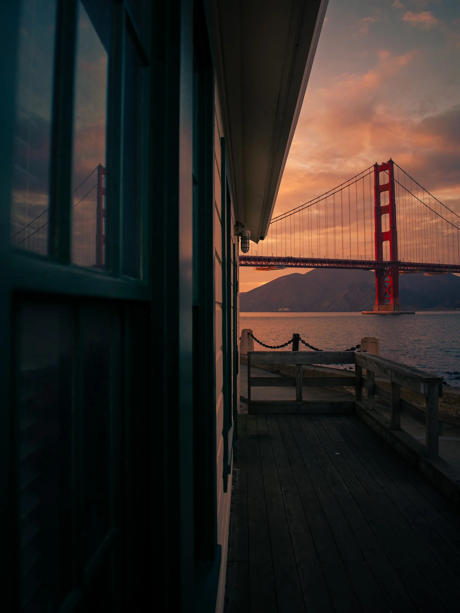 Sunset view of the Golden Gate Bridge seen from a wooden deck next to a house, with reflections in a window on the left.