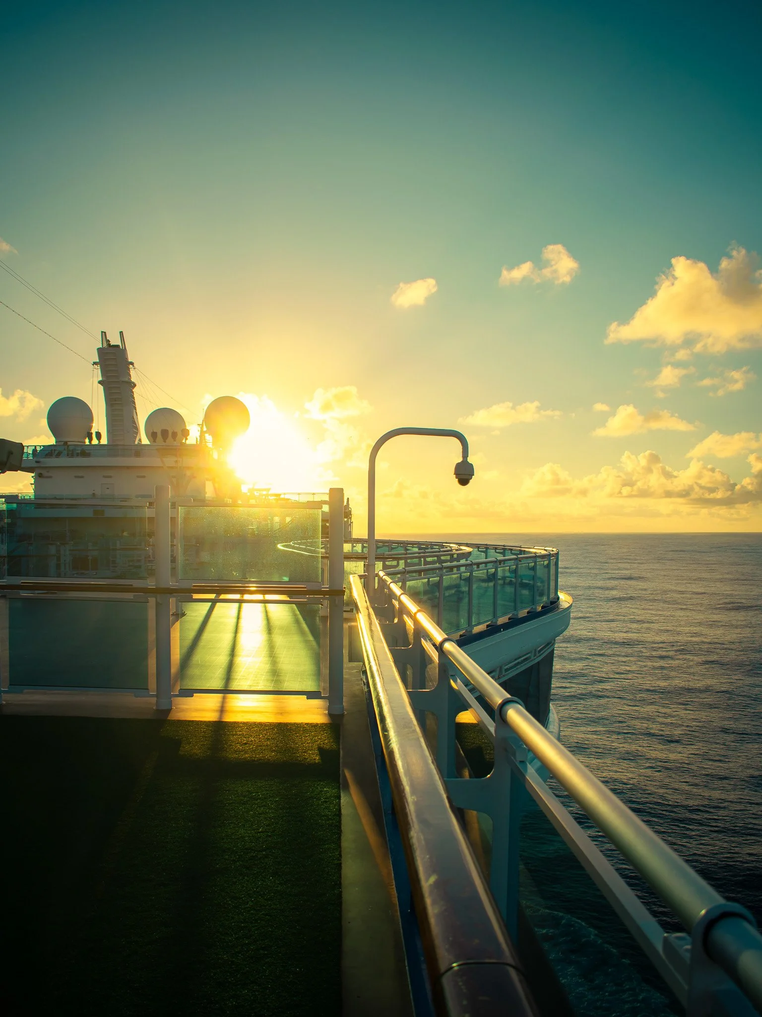 The deck of a cruise ship at sunset, showing a walkway with metal railings, satellite antennas, and a surveillance camera, with the ocean and a partly cloudy sky in the background.