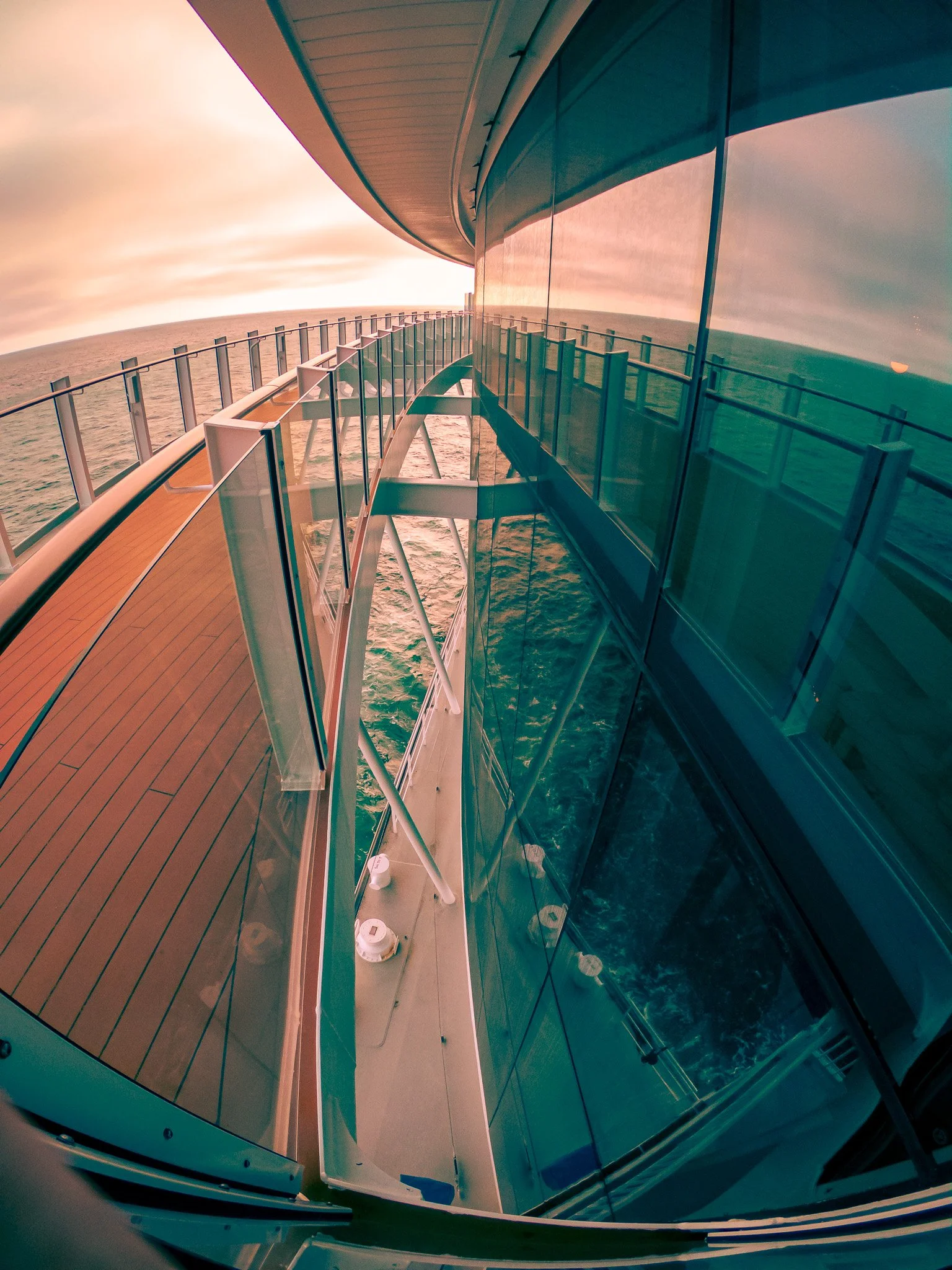 View of the exterior of a cruise ship with a curved glass wall and a wooden deck, overlooking the ocean during sunset.