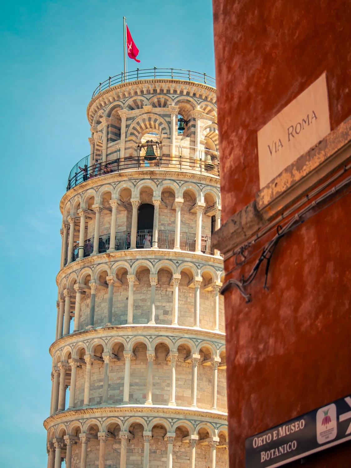 The Leaning Tower of Pisa against a blue sky with a red wall and a sign reading "VIA ROMA" in the foreground.