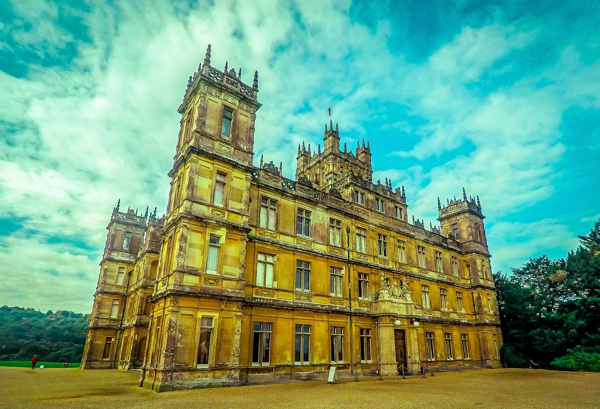 Downton Abbey (Highclere Clastle) surrounded by a grassy area and trees under a partly cloudy sky.