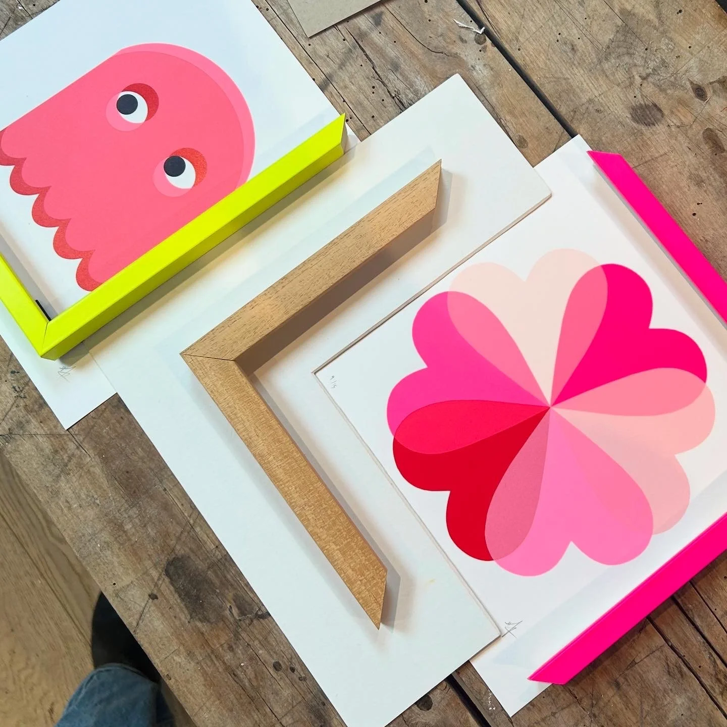 Colorful artwork featuring a pink cartoon-like octopus and a pink and red geometric heart design, with different colored frames and paper around them on a wooden table. Neon coloured Frames and Spacers
