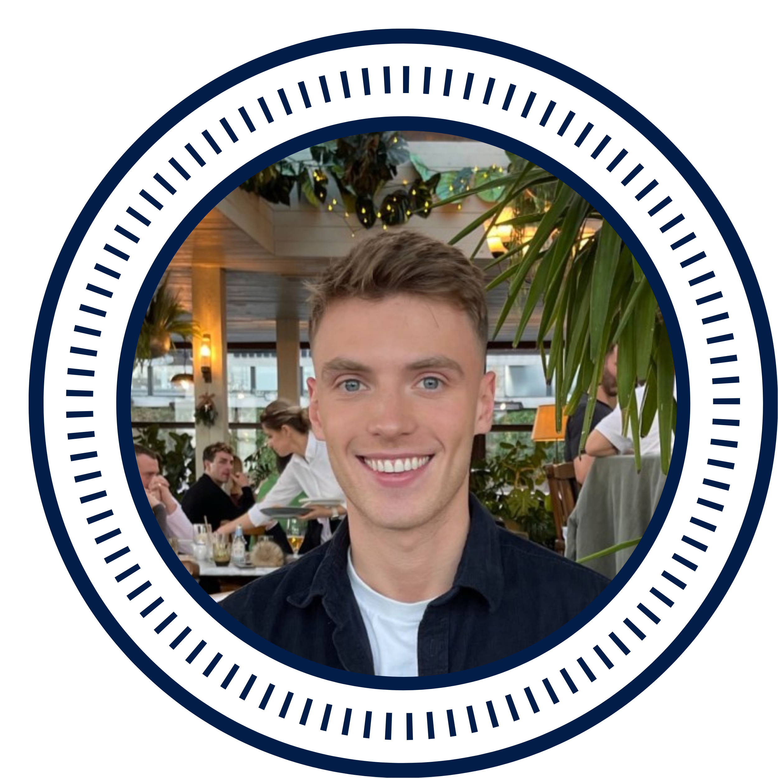 Young man with short light brown hair and blue eyes smiling in a restaurant with greenery and people in the background.