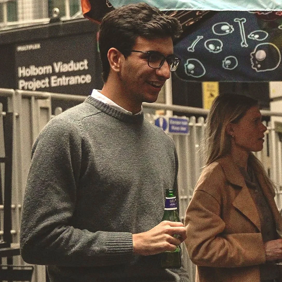 A man with dark hair, glasses, and a gray sweater holding a beer bottle, standing next to a woman with long hair and a brown coat at an outdoor event under a table decorated with skull and bones designs, near the Hobson Valley Project Entrance sign.